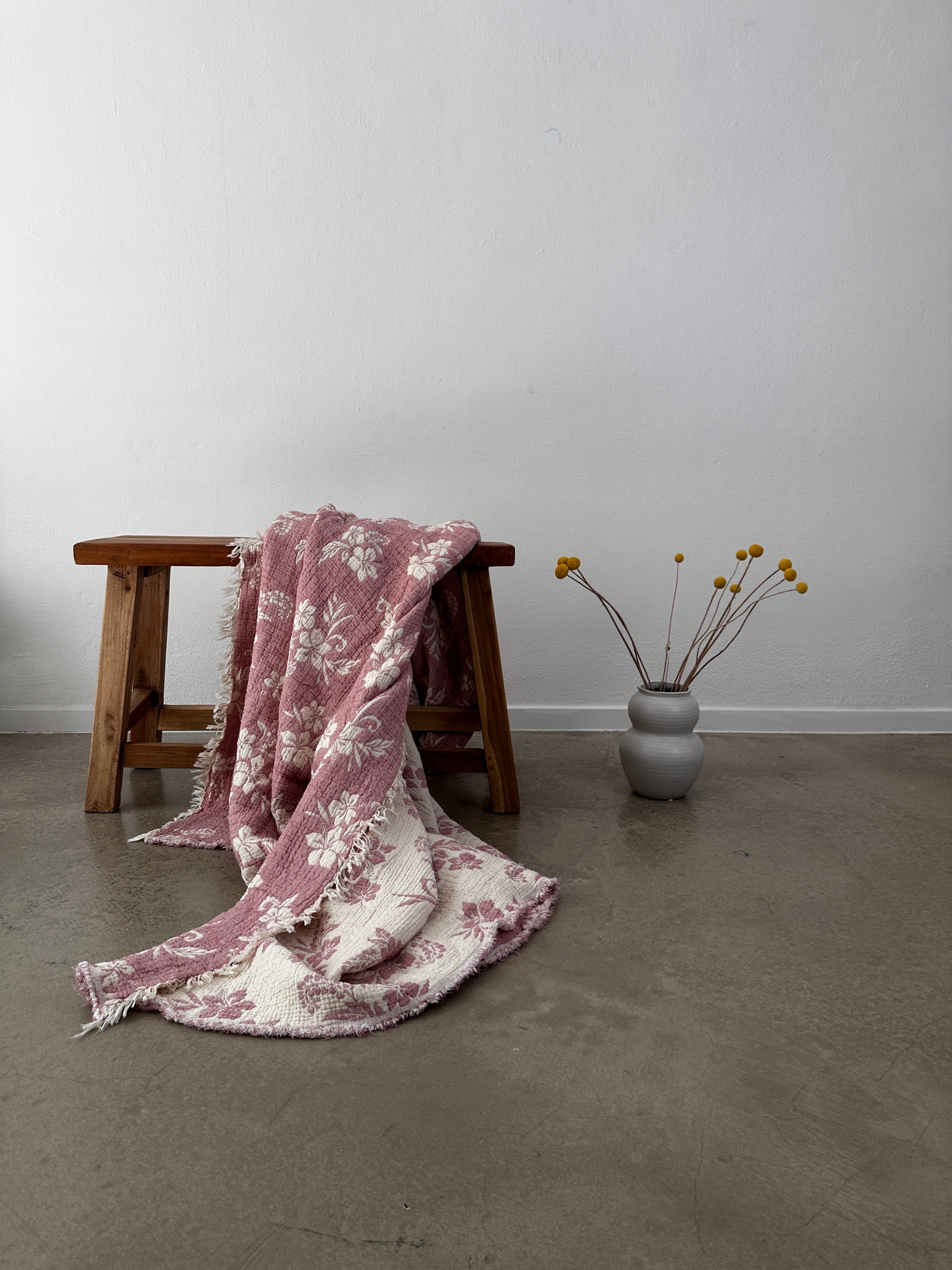 Eine Holzbank zeigt die LEY! Musselin Decke - ROSE in Altrosa-Weiß (2,20 x 2,50 m), elegant drapiert auf Betonboden. Eine graue Vase mit gelben Billiardkugeln ergänzt den Boho-Stil vor einer schlichten weißen Wand.