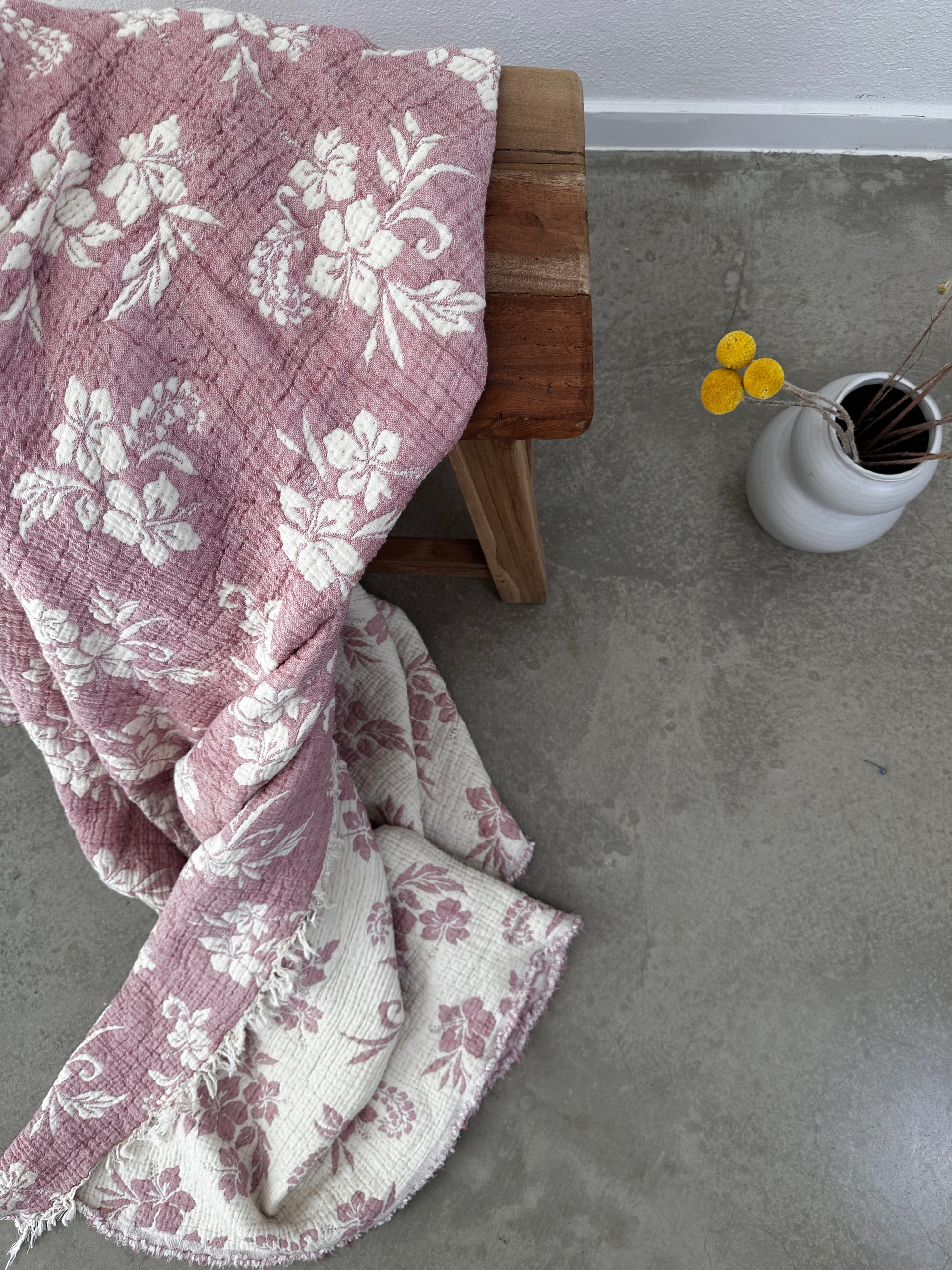 Die LEY! Musselin Decke - ROSE in Altrosa-Weiß (2,20 x 2,50 m), drapiert über einem Holzhocker auf einem Betonboden, mit gelben Ballblüten in einer weißen Vase daneben, verleiht der Szene charmantes Boho-Design-Flair.