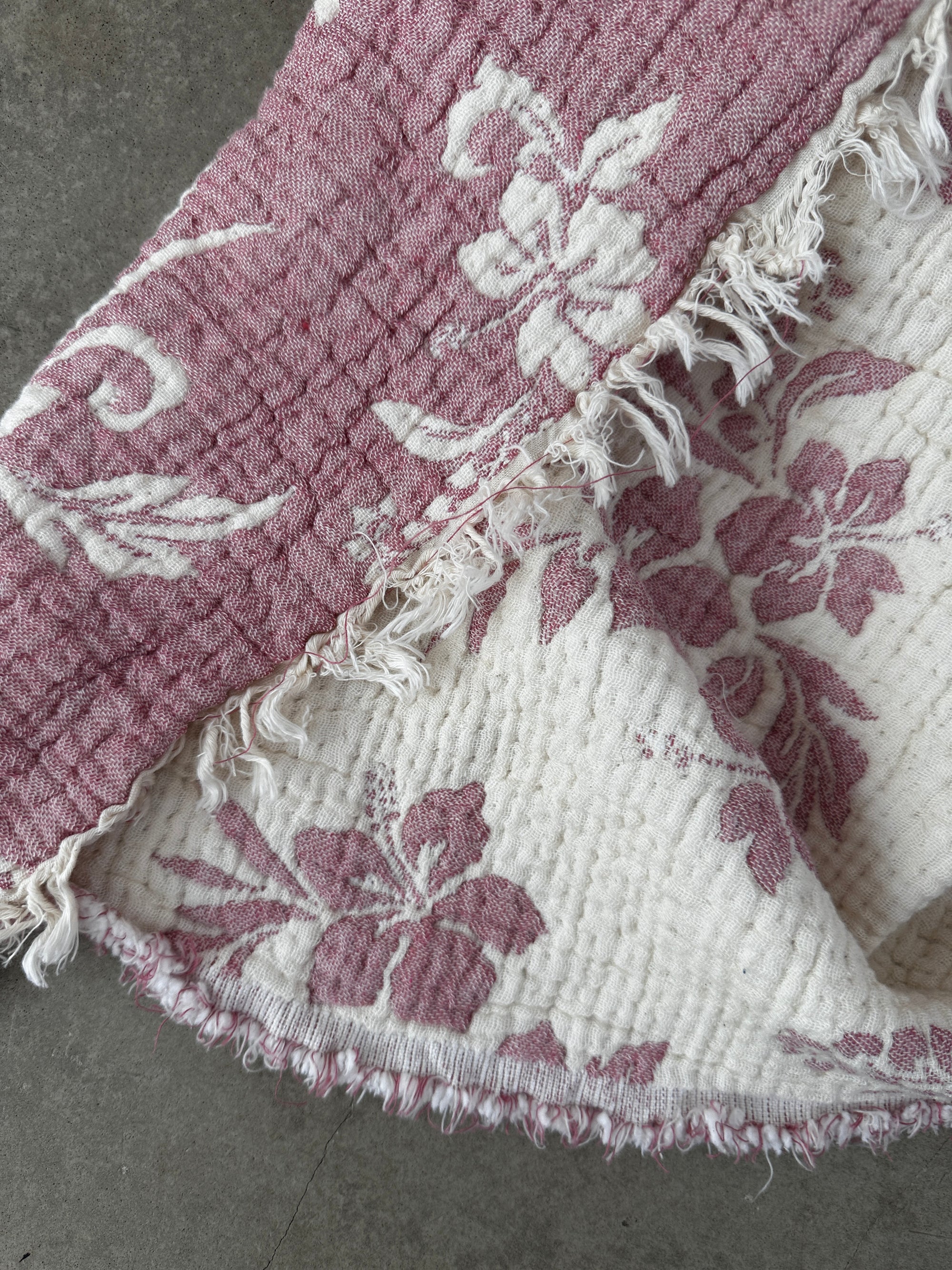 Eine Nahaufnahme der LEY! Musselin Decke - ROSE in Altrosa-Weiß (2,20 x 2,50 m) zeigt ihr strukturiertes florales Muster mit rosa und weißen Hibiskusblüten, ausgefransten Kanten und gefaltetem Stoff, der beide Seiten auf einer grauen Oberfläche zeigt.