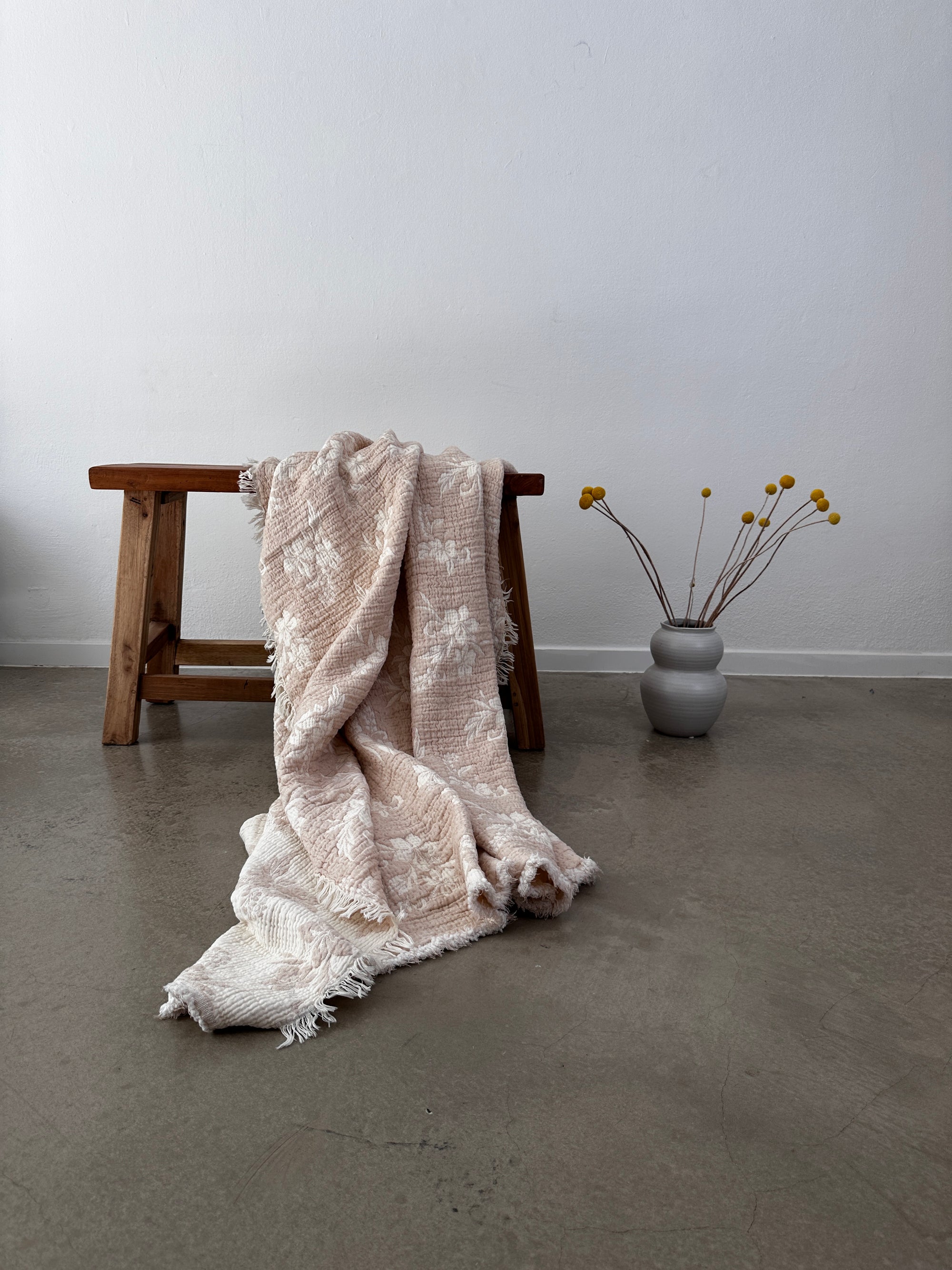 Auf einer Holzbank ist die LEY! Musselin Decke - ROSE in Altrosa-Weiß (2,20 x 2,50 m) darüber drapiert. Daneben steht eine graue Vase mit gelben Bällen, die den Boho-Stil an die schlichte weiße Wand und den Betonboden bringt.