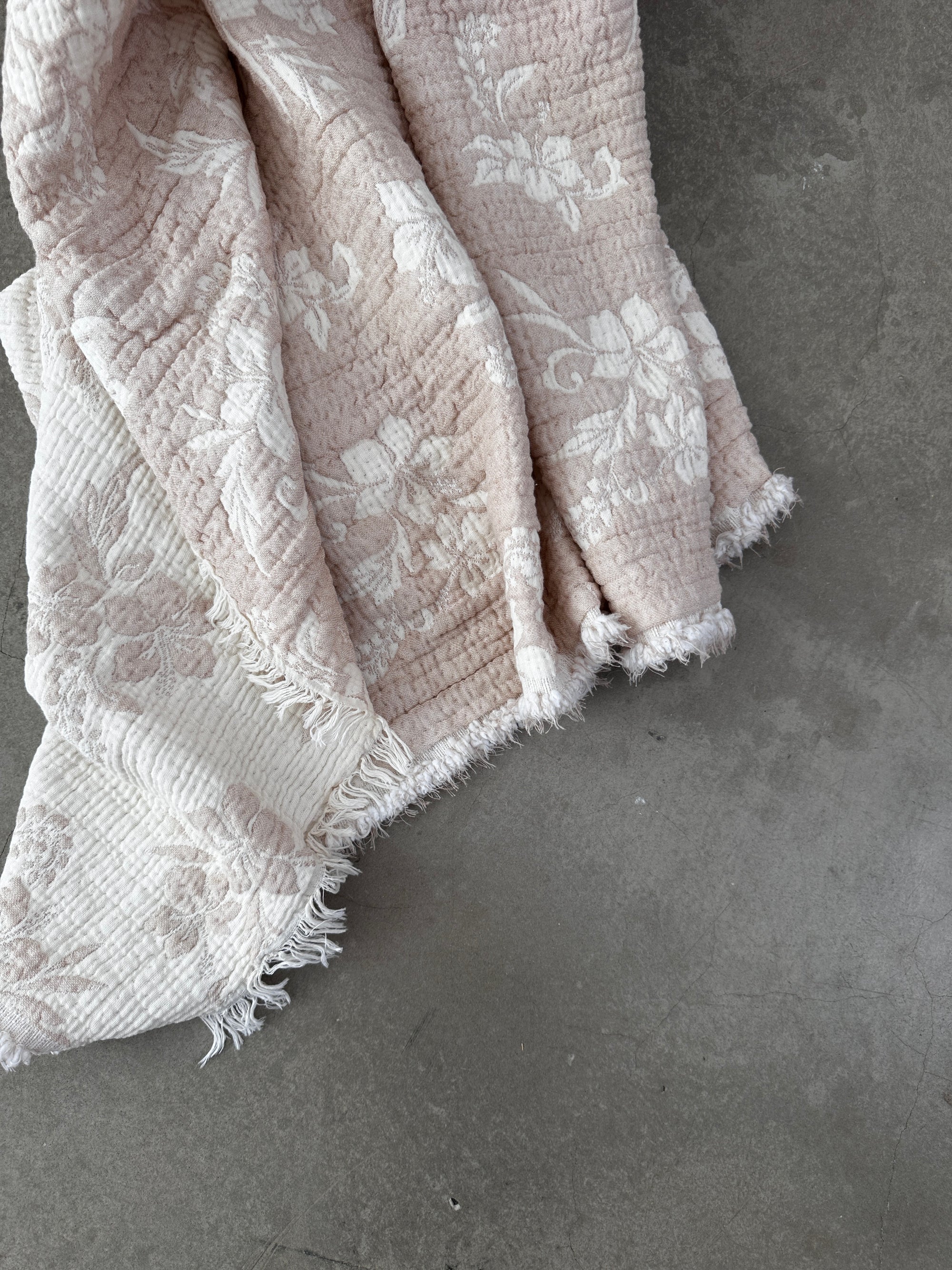 Die LEY! Musselin Decke - ROSE in Beige-Weiß (2,20 x 2,50 m) zeigt ein zartes, florales Boho-Muster mit ausgefransten Rändern, elegant drapiert auf einer glatten grauen Oberfläche.