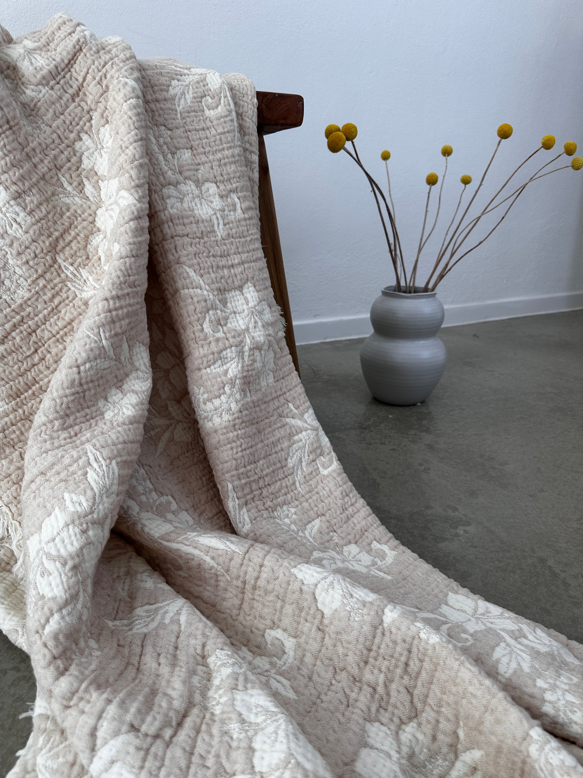 Die Musselin Decke - ROSE von LEY! in Beige-Weiß (2,20 x 2,50 m) mit weißen Blumenmustern liegt im Boho-Stil über einem Holzstuhl neben einer grauen Vase mit gelben Billy Balls auf Zementboden vor weißer Wand.
