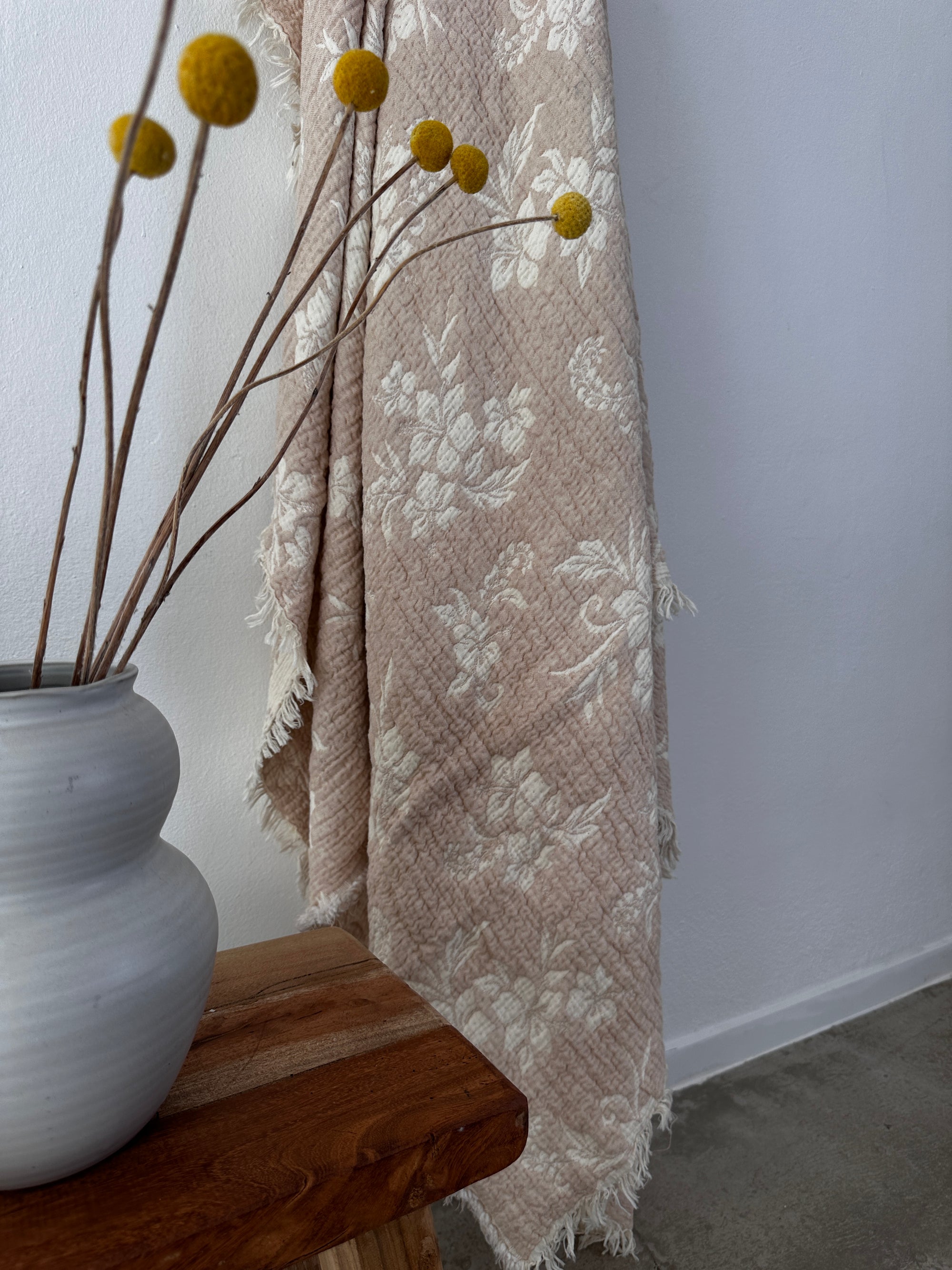 Eine beige-weiße Musselin Decke - ROSE by LEY! (2,20 x 2,50 m) mit Blumendruck und Fransen hängt neben einer Holzbank, auf der eine graue Vase mit gelben Bällen und getrockneten Zweigen für einen weichen Boho-Look sorgt.