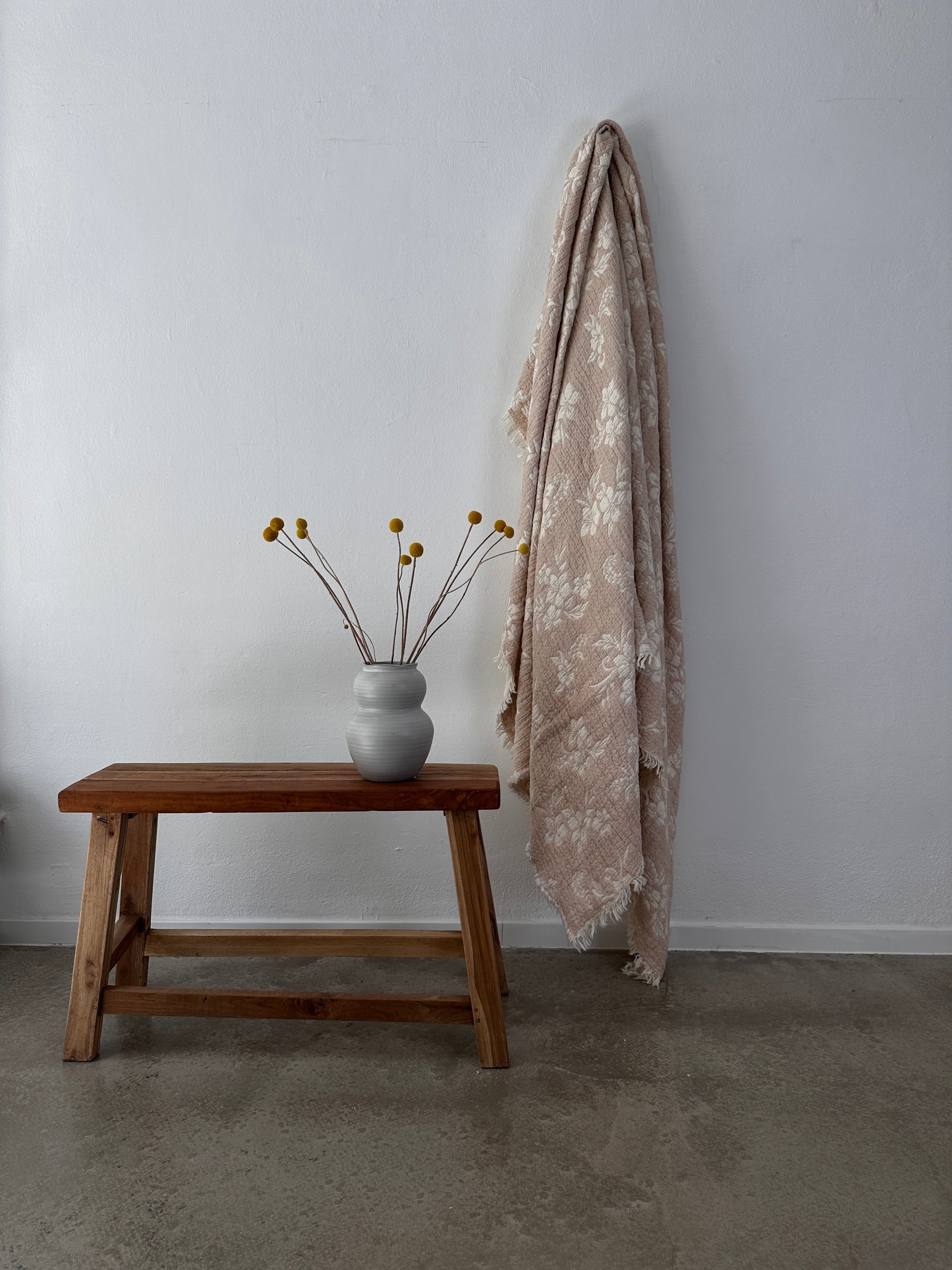Eine Holzbank mit einer grauen Vase, in der gelbe Billy-Button-Blumen stehen, steht neben der LEY! Musselin Decke - ROSE in Beige-Weiß (2,20 x 2,50 m), drapiert im Boho-Stil an einer weißen Wand über einem polierten Betonboden.