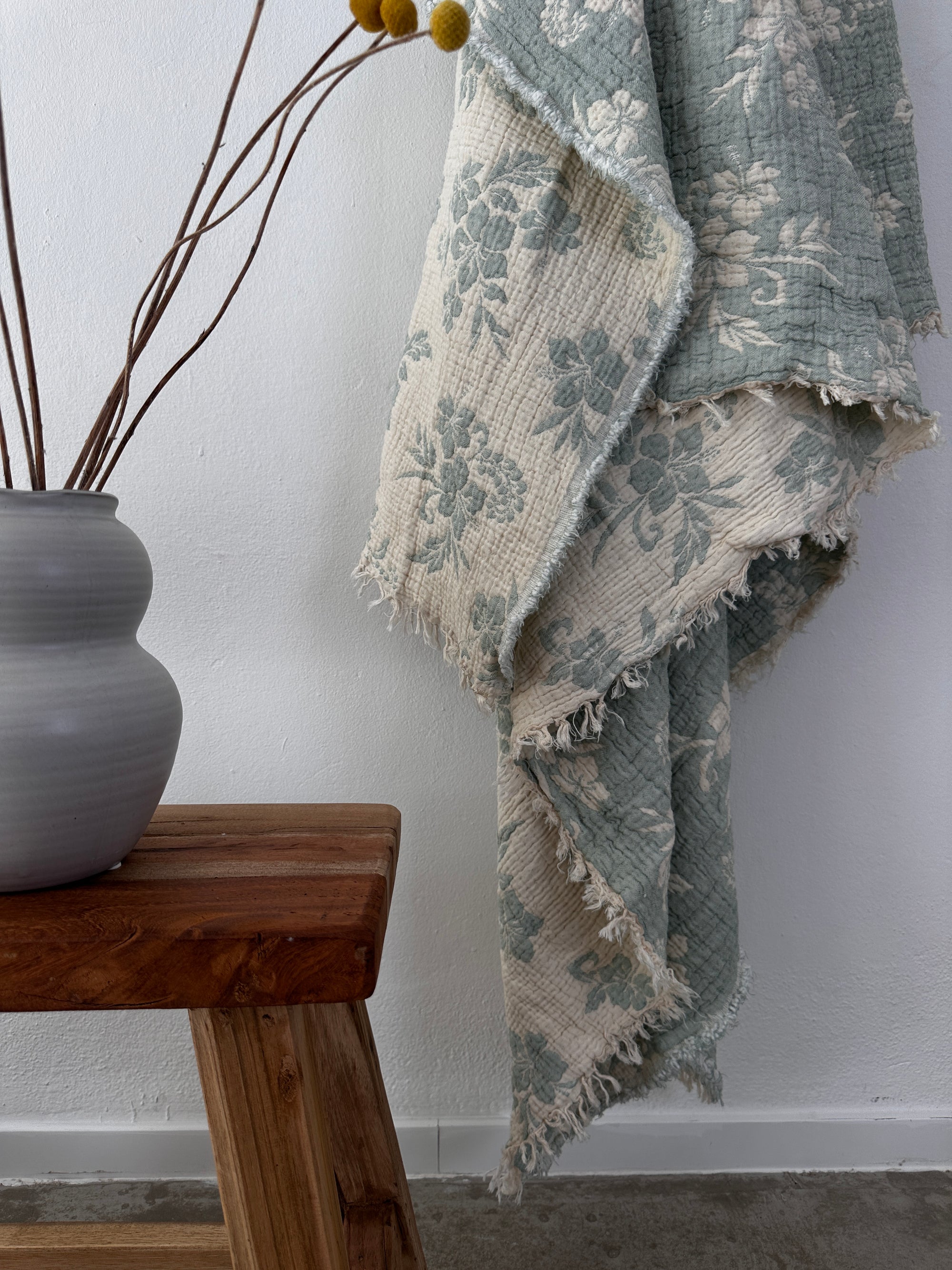 Die LEY! Musselin Decke - ROSE in hellblau-weiß (2,20 x 2,50 m) setzt mit ihrem floralen Muster und den ausgefransten Rändern einen gemütlichen Boho-Akzent, gestylt neben einer grauen Keramikvase mit getrockneten Stängeln auf einer Holzbank vor einer weißen Wand.