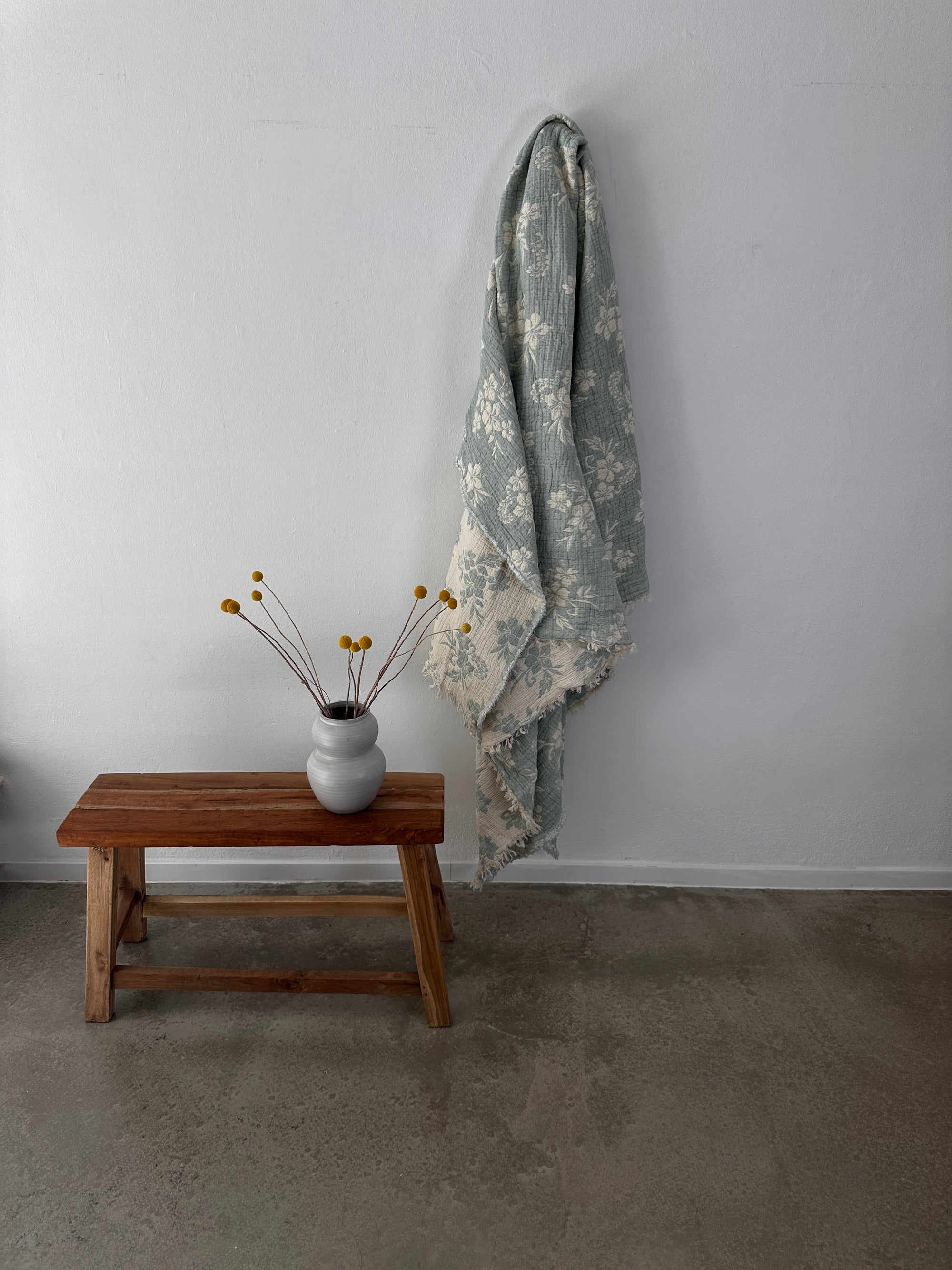 Eine Holzbank mit einer weißen Vase mit gelben Billyball-Blüten steht neben der LEY! Musselin Decke - ROSE in hellblau-weiß (2,20 x 2,50 m), deren weiches Boho-Design an einer weißen Wand über einem Betonboden drapiert ist.