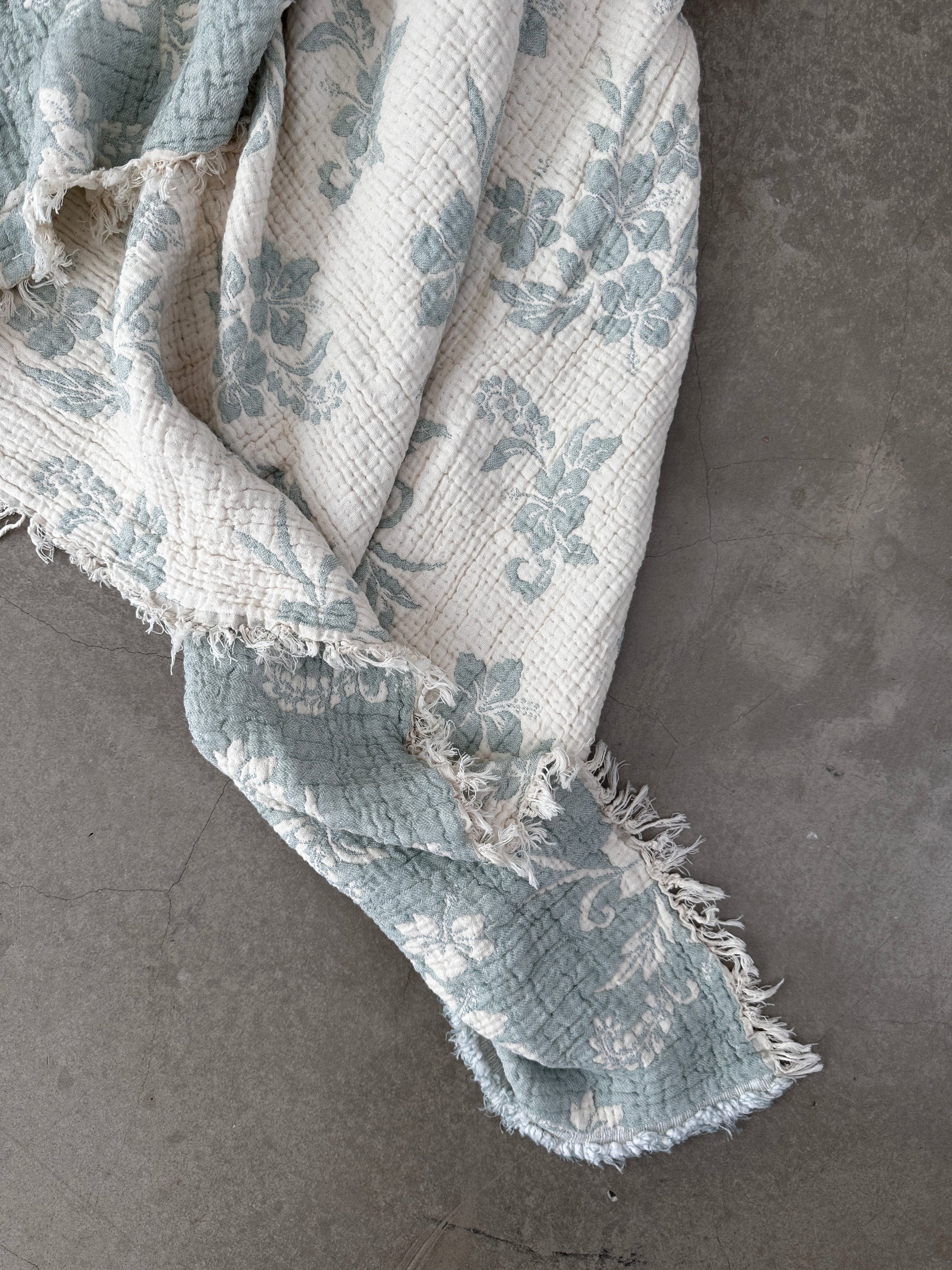 Die LEY! Musselin Decke - ROSE in Hellblau-Weiß (2,20 x 2,50 m) bringt mit ihrer gesteppten Struktur und den ausgefransten Kanten Boho-Design-Charme in Ihren Raum, schön drapiert auf einem schlichten grauen Betonboden.