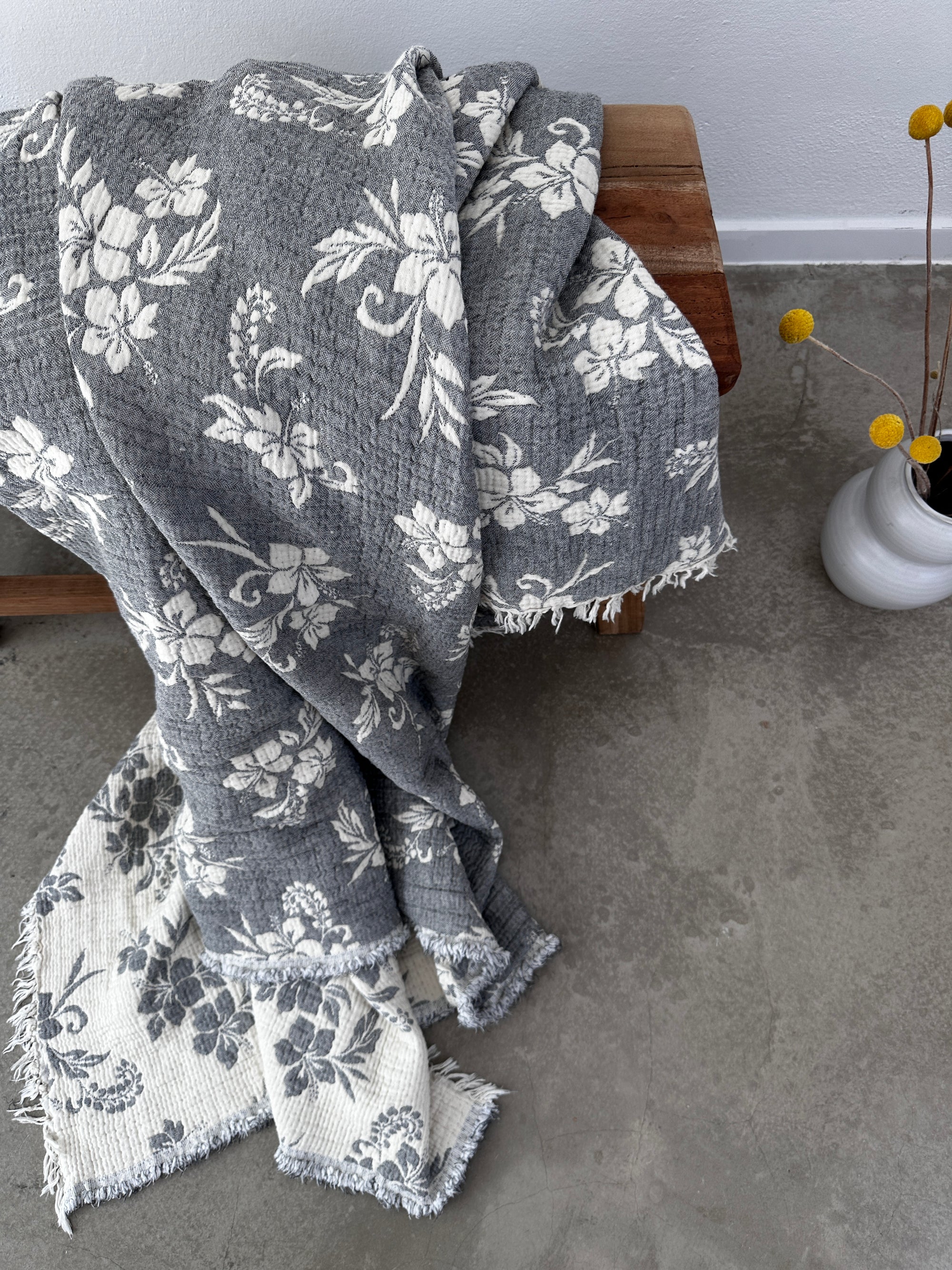 Die Musselin Decke - ROSE von LEY! in Grau-Weiß (2,20 x 2,50 m) mit Boho-Design und Fransen liegt auf einer Holzbank; daneben eine weiße Vase mit gelben Billy Ball Blumen auf poliertem Betonboden.