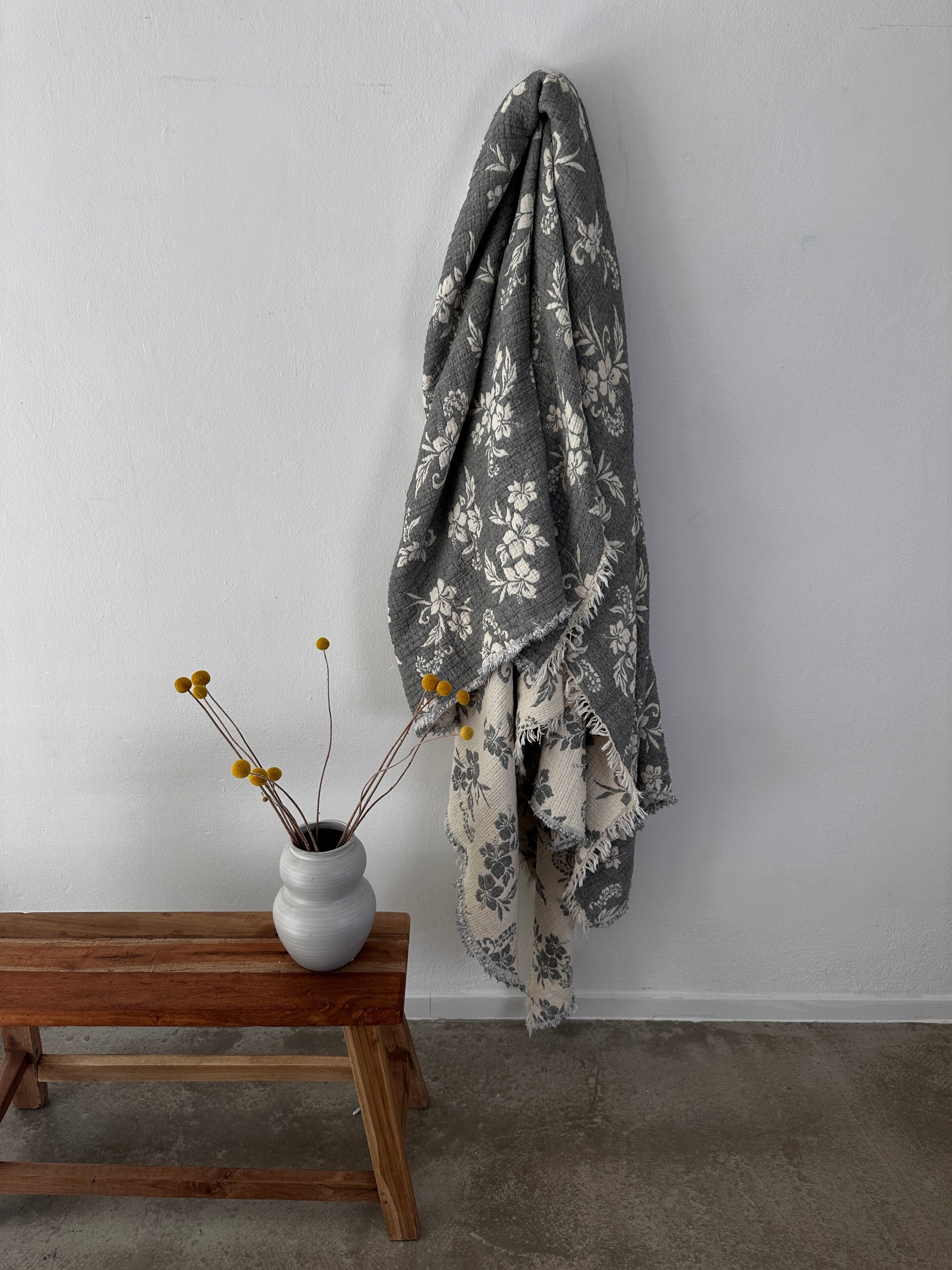 Die Musselin Decke - ROSE von LEY! in Grau-Weiß (2,20 x 2,50 m) mit Boho-Blumenmuster hängt an einer weißen Wand über einer Holzbank. Auf der Bank steht eine Vase mit gelben Billy Balls, der Boden ist grauer Beton.