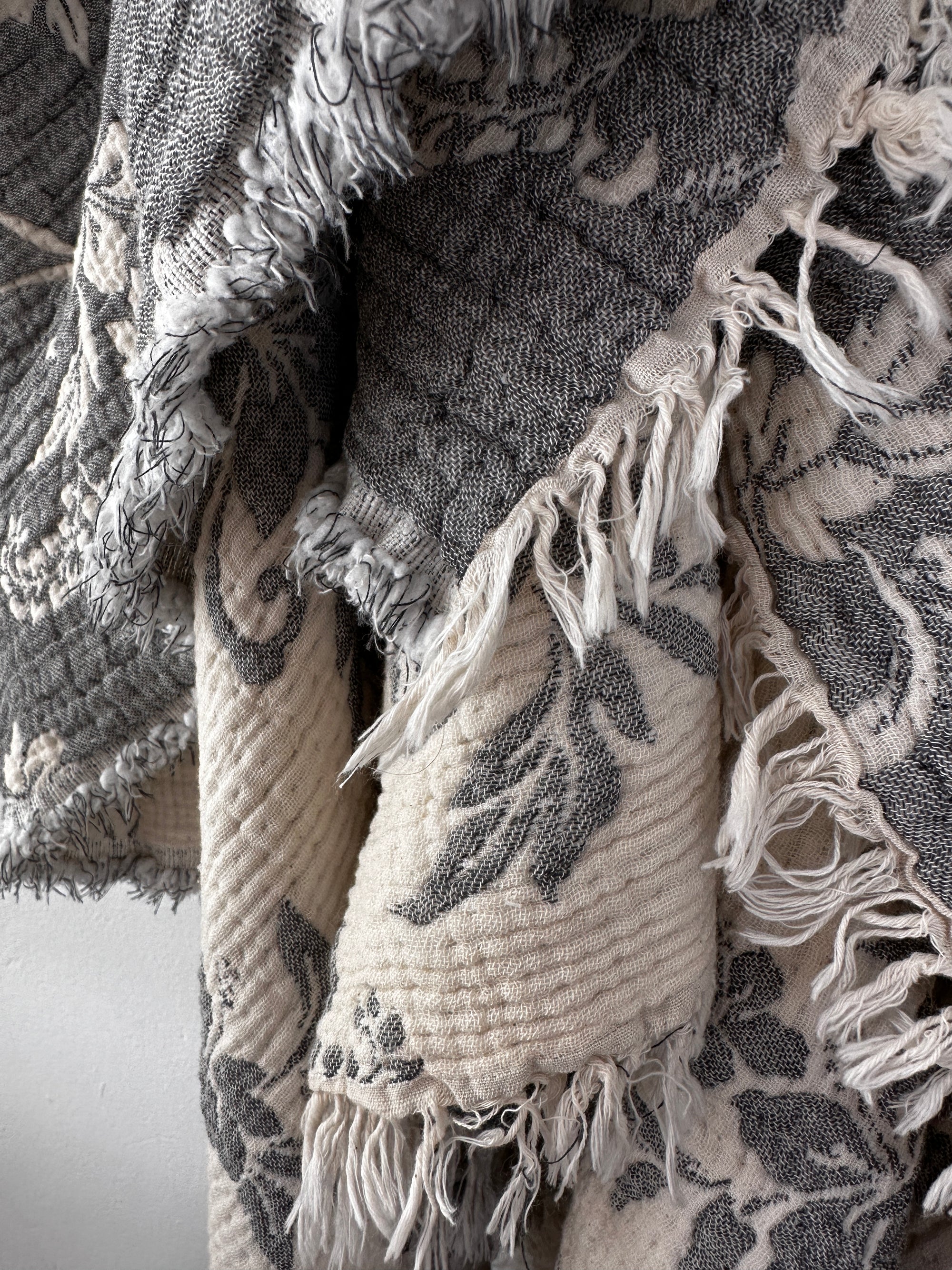 Nahaufnahme der LEY! Musselin Decke - ROSE in Grau-Weiß (2,20 x 2,50 m) mit floralem Muster, ausgefransten Kanten und Boho-Design. Das weiche, mehrlagige Material sorgt für einen gemütlichen, rustikalen Look.