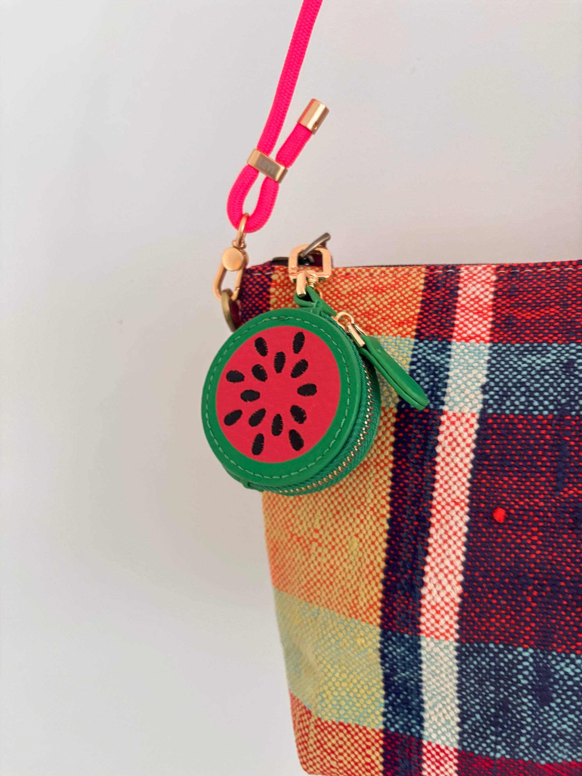 Eine Nahaufnahme des LEY! Schlüsselanhänger - Melone, einem runden Münzbeutel aus grünem Kunstleder mit roter, mit Samen verzierter Mitte und rosafarbenem Riemen, der an einer karierten Stofftasche befestigt ist.