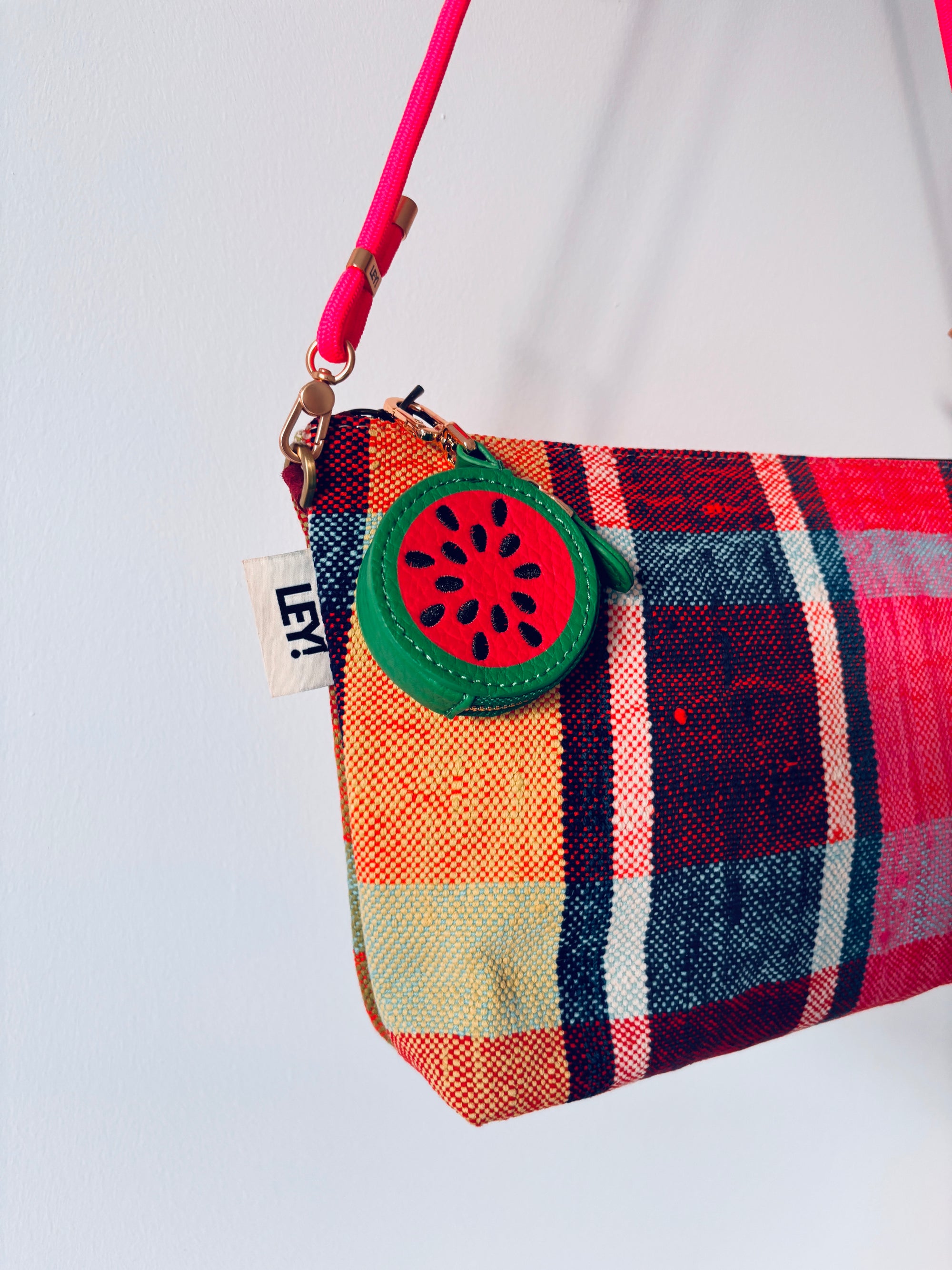 Eine bunt karierte LEY! Mini Etui Kunstleder Handtasche mit einem Schlüsselanhänger - Melone und einem rosafarbenen Riemen, der vor einem weißen Hintergrund hängt.