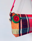 Eine bunt karierte LEY! Mini Etui Kunstleder Handtasche mit einem Schlüsselanhänger - Melone und einem rosafarbenen Riemen, der vor einem weißen Hintergrund hängt.