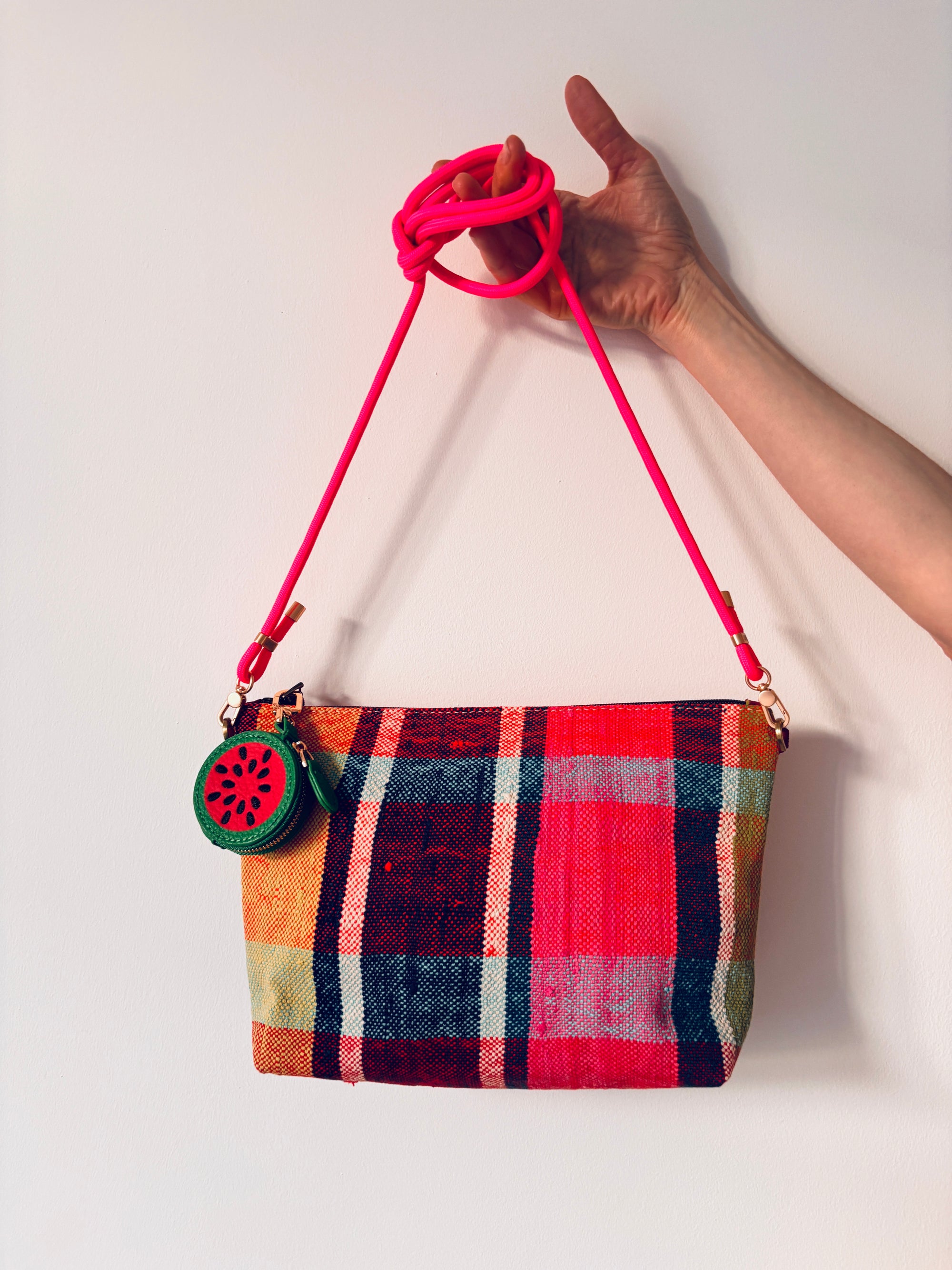 Eine Hand hält eine rot-rosa karierte Mini Etui Kunstleder Handtasche mit einem hellrosa Riemen und dem LEY! Schlüsselanhänger - Melone, vor einem schlichten weißen Hintergrund.