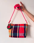 Eine Hand hält eine rot-rosa karierte Mini Etui Kunstleder Handtasche mit einem hellrosa Riemen und dem LEY! Schlüsselanhänger - Melone, vor einem schlichten weißen Hintergrund.