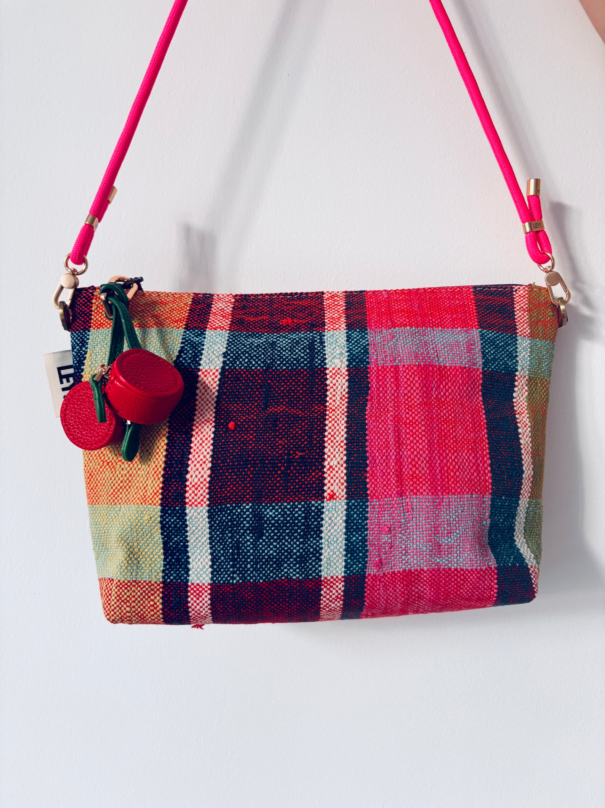 Eine rechteckige LEY! Handtasche mit einem lebhaften Karomuster in Rot, Pink, Grün, Blau und Gelb, mit einem pinkfarbenen Riemen und zwei Schlüsselanhängern - Kirsche, die am Reißverschluss hängen.