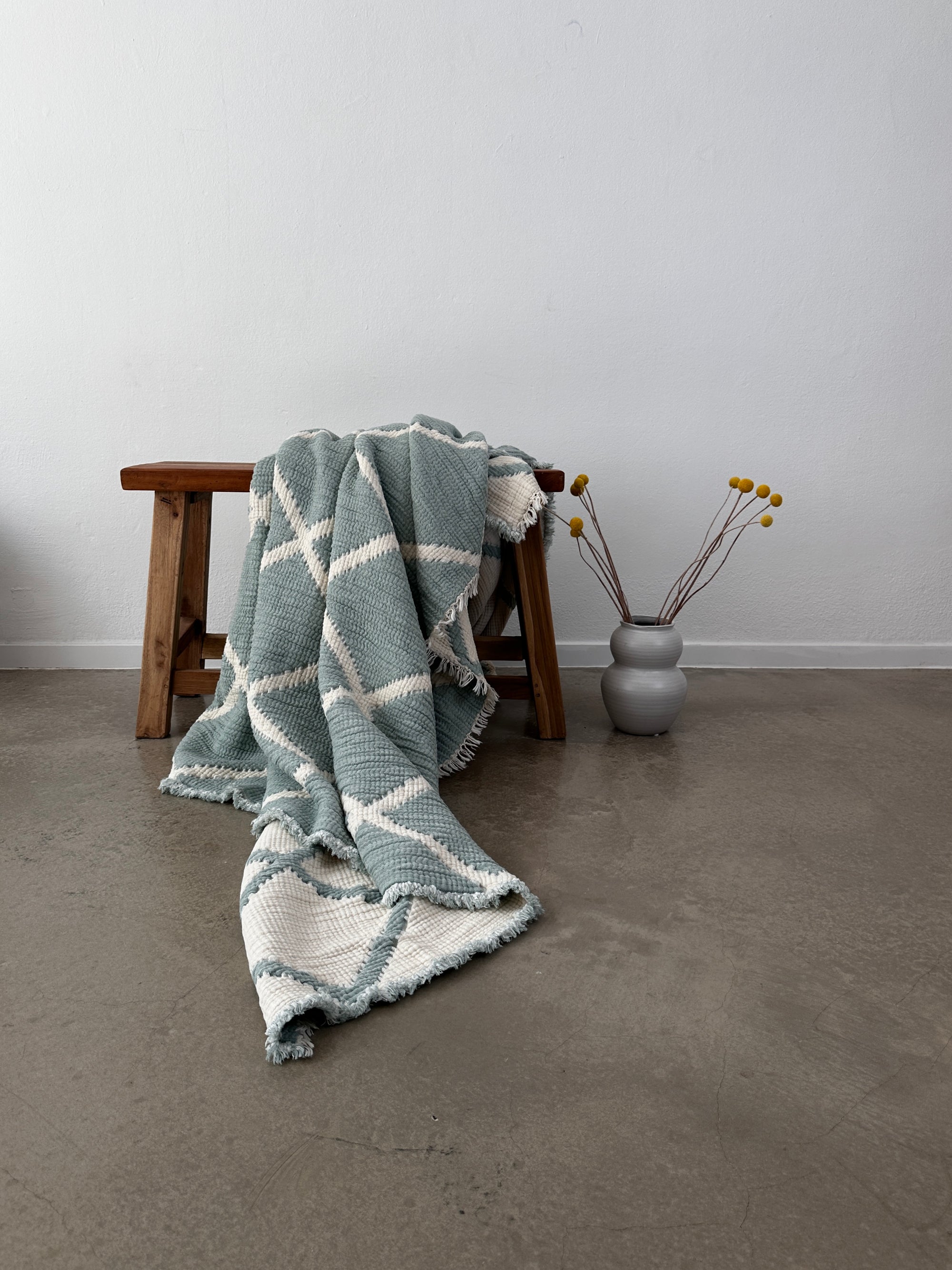 Eine Holzbank mit einer LEY! Musselin Decke in Salbei-Weiß (2,20 x 2,50 m) ist darüber drapiert, neben einer grauen Vase mit gelben Billy Ball Blumen auf poliertem Beton vor einer weißen Wand.