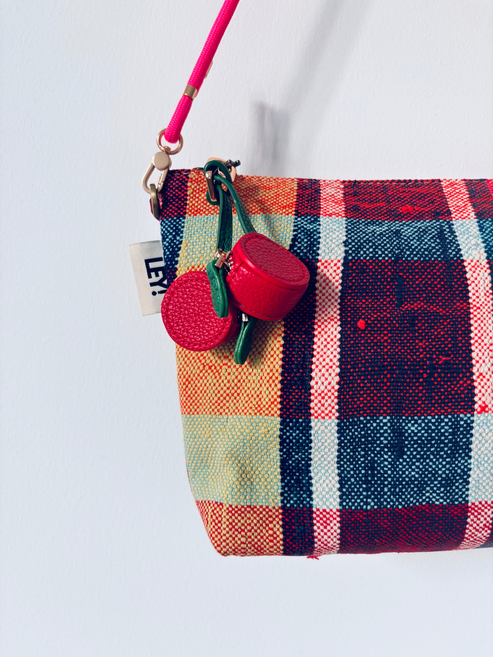 An der Wand hängt eine bunt karierte Mini-Etui-Kunstleder-Handtasche mit rosafarbenem Riemen, die mit dem LEY! Schlüsselanhänger - Kirsche und einem kleinen weißen Anhänger mit teilweise sichtbarer schwarzer Schrift.