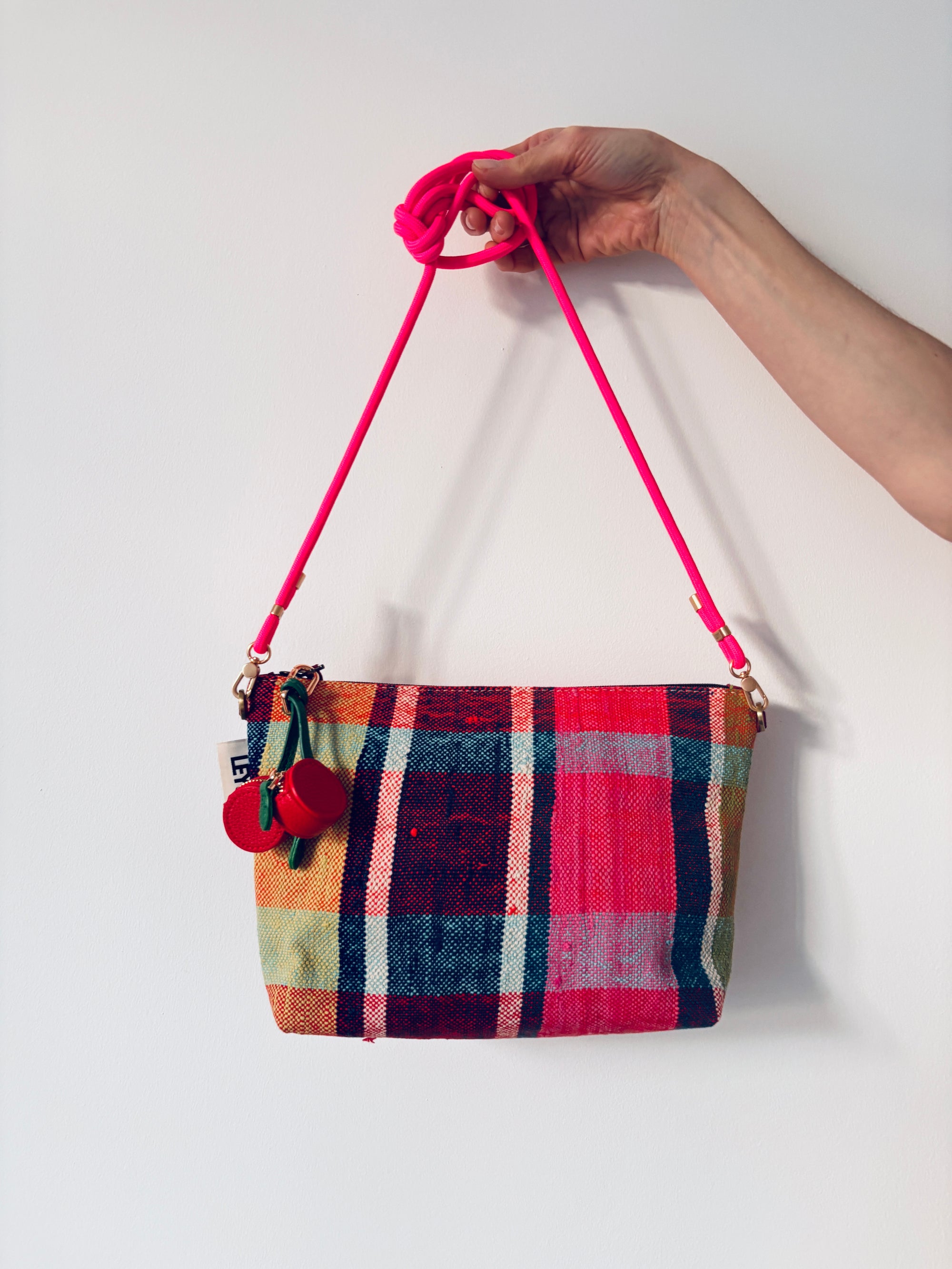 Eine Hand hält eine bunt karierte Handtasche mit einem leuchtend rosa Riemen und einer LEY! Schlüsselanhänger - Kirsche vor einem schlichten weißen Hintergrund.