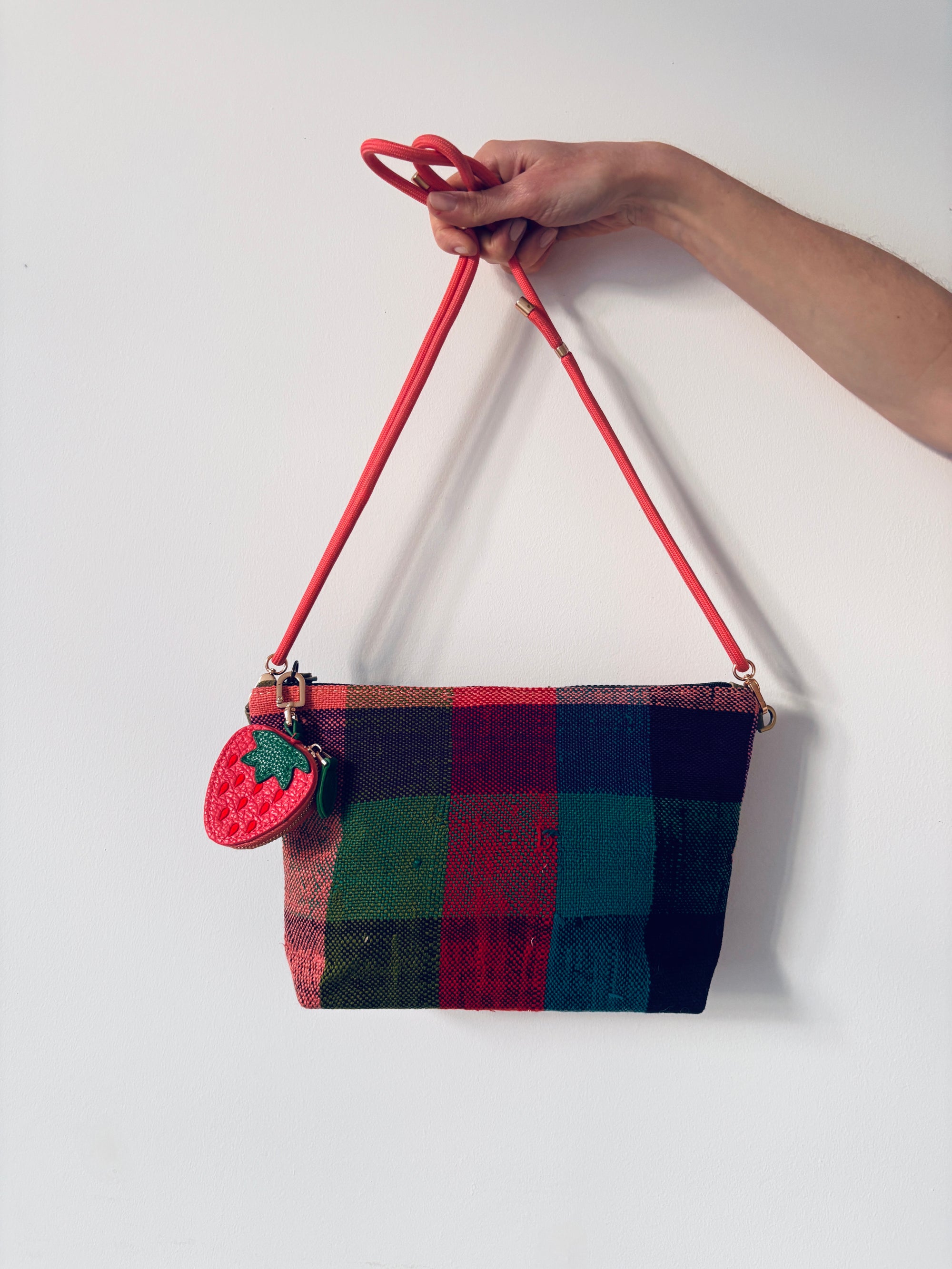 Eine Hand hält eine karierte Umhängetasche mit roten, grünen und schwarzen Quadraten, die mit einem roten Riemen und einem LEY! Schlüsselanhänger - Erdbeere aus Mini Etui Kunstleder am Griff vor weißem Hintergrund.