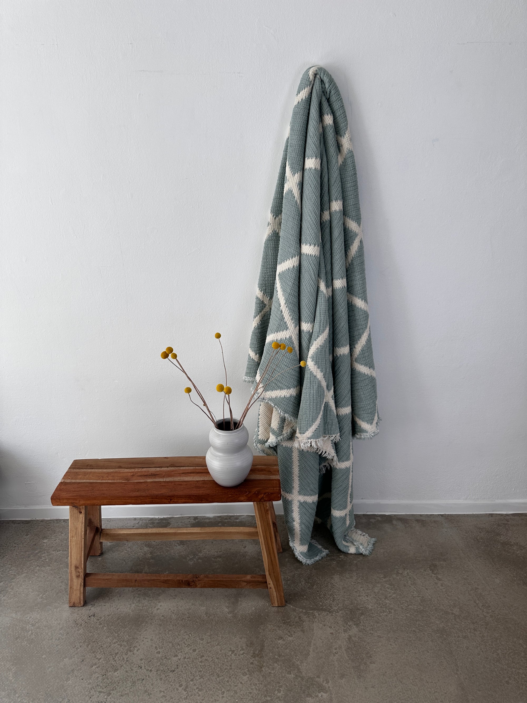 Die Musselin Decke - Salbei-Weiß von LEY! (2,20 x 2,50 m) im Boho-Design mit geometrischem Muster hängt vor einer weißen Wand neben einer Holzbank mit Vase und gelben Blumen auf einem Betonboden.