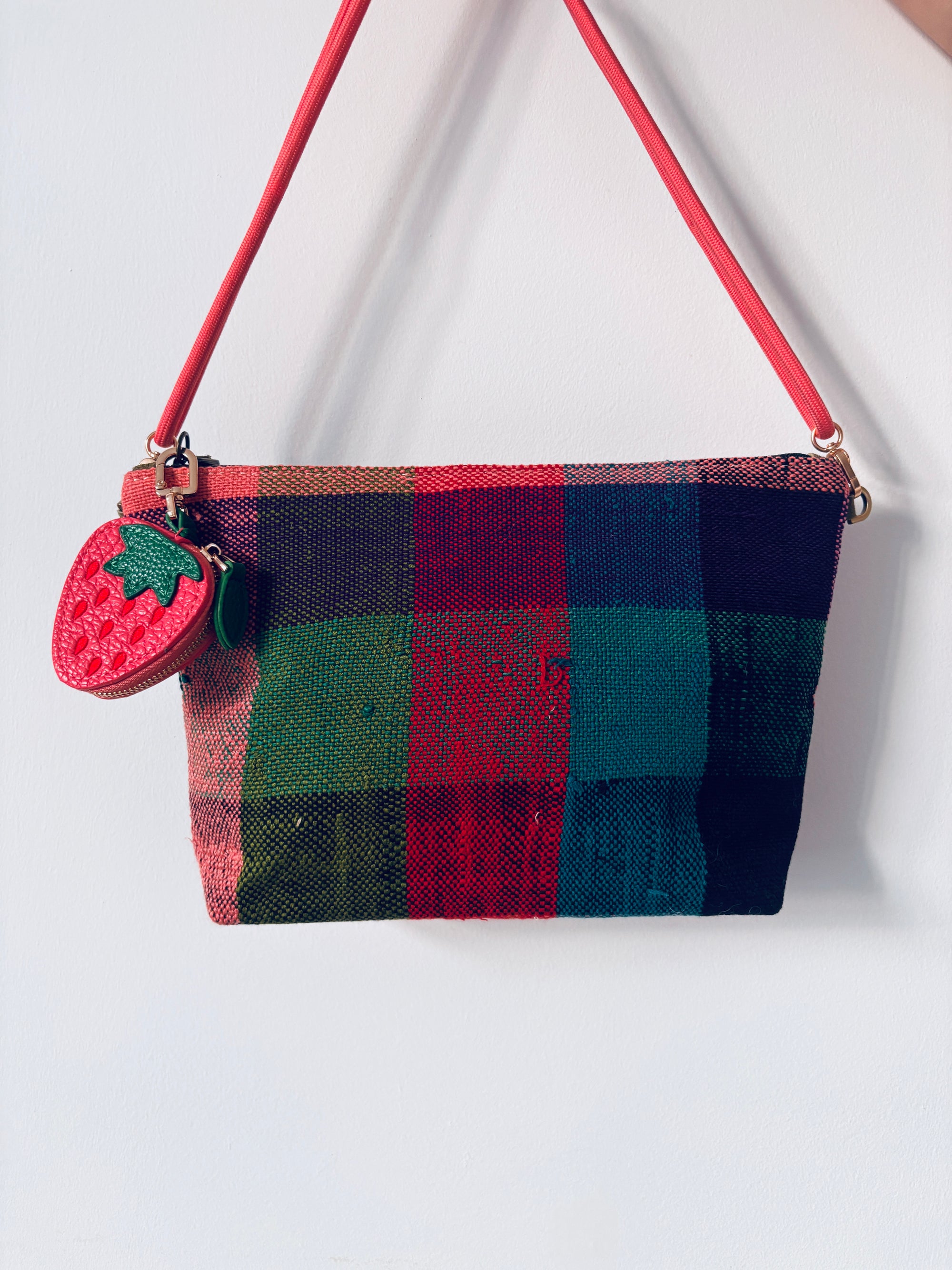 Eine bunt karierte Handtasche mit rotem Riemen und einem LEY! Schlüsselanhänger - Erdbeere hängt an einer schlichten weißen Wand.
