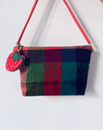 Eine bunt karierte Handtasche mit rotem Riemen und einem LEY! Schlüsselanhänger - Erdbeere hängt an einer schlichten weißen Wand.