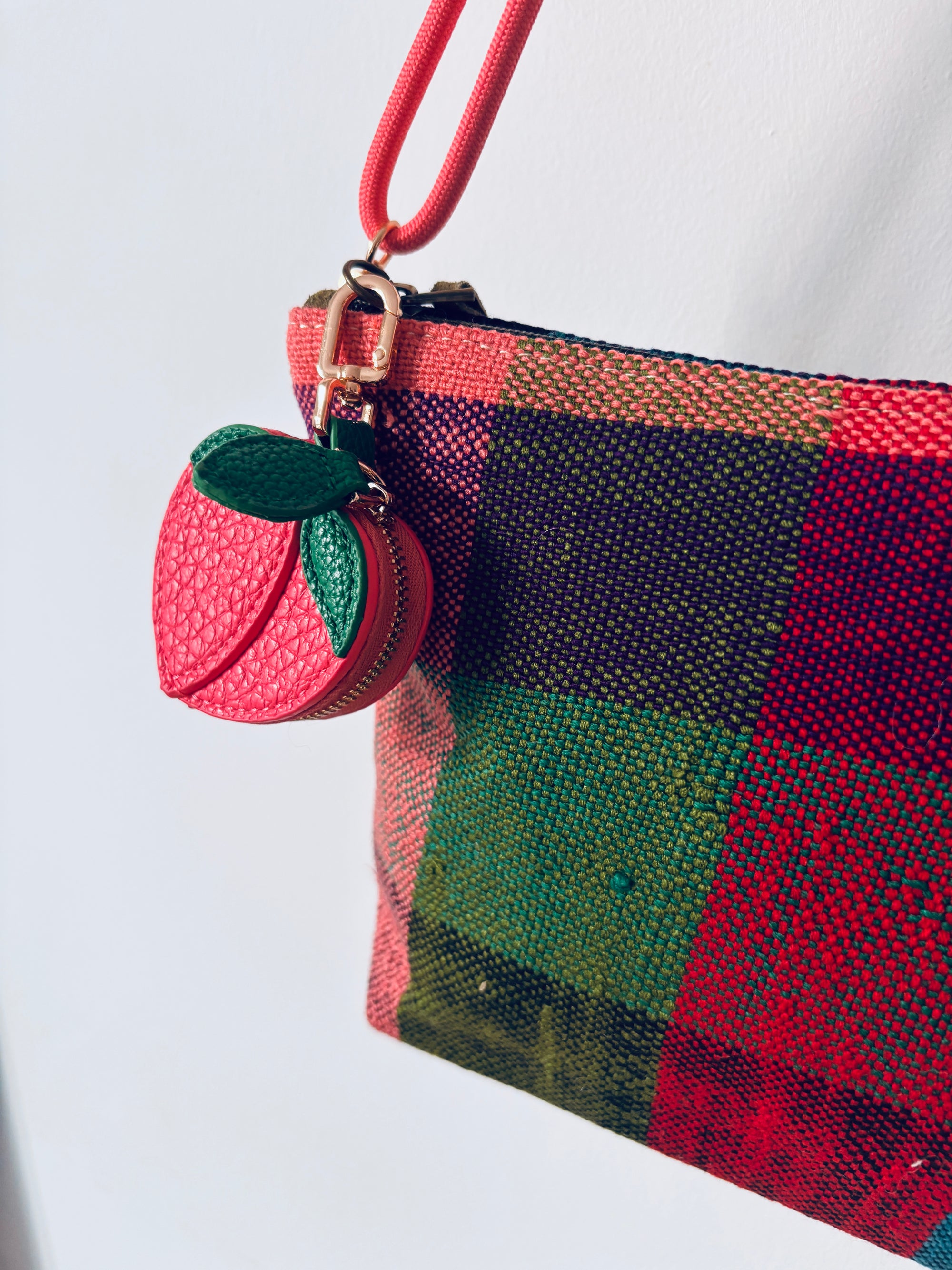 Eine Nahaufnahme eines lebhaft karierten Beutels mit einem rot-grün-schwarzen Karomuster. Der Reißverschluss ist mit dem Schlüsselanhänger - Pfirsich von LEY! verziert, was dieses Mini-Etui aus Kunstleder sowohl praktisch als auch stilvoll macht.