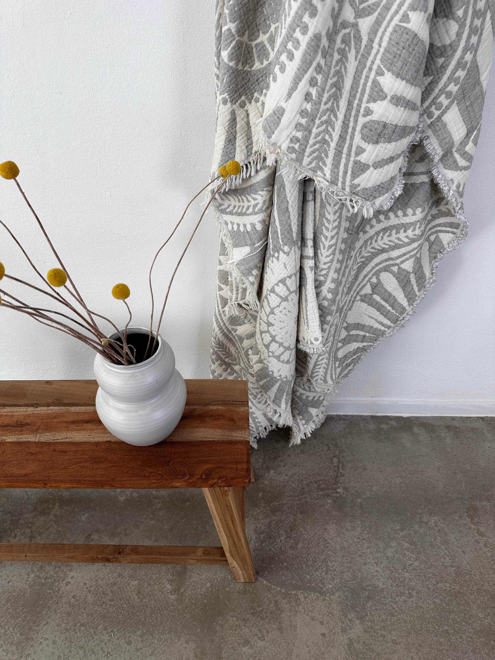 Eine weiße Vase mit gelben Billyknöpfen steht auf einer Holzbank neben der LEY! CALMA Musselin Decke in hellgrau (2,20 x 2,30 m), drapiert an einer weißen Wand auf einem Betonboden.