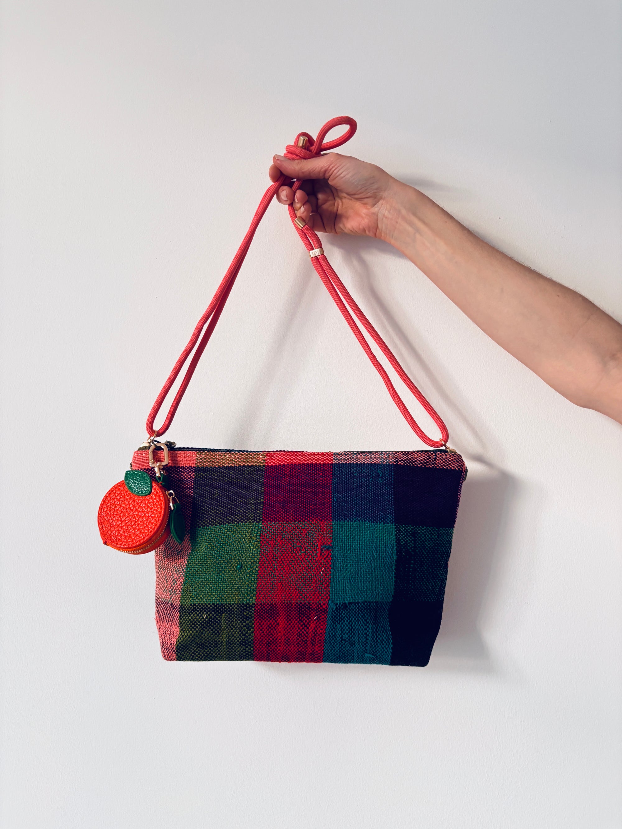 Eine Hand hält einen LEY! Schlüsselanhänger - Etui - Orange und eine karierte Handtasche mit rotem Riemen vor weißem Hintergrund.