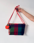 Eine Hand hält einen LEY! Schlüsselanhänger - Etui - Orange und eine karierte Handtasche mit rotem Riemen vor weißem Hintergrund.
