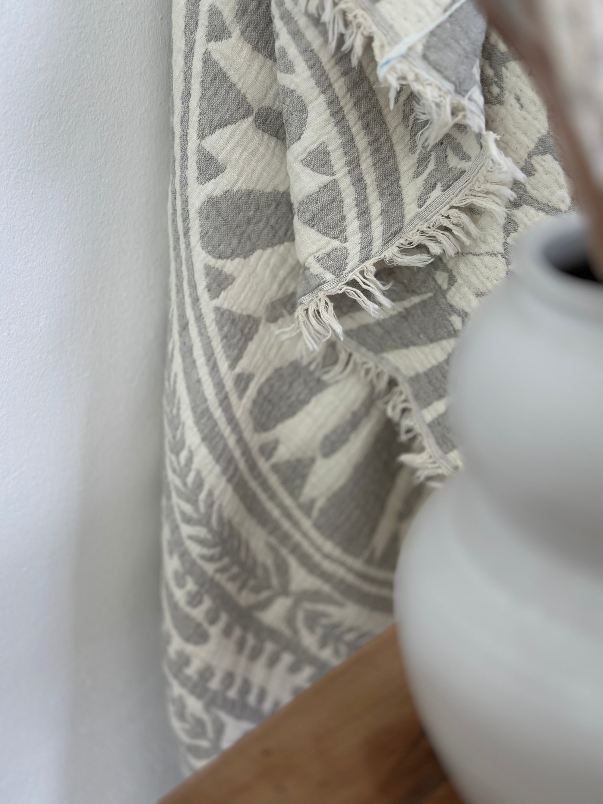 Eine Nahaufnahme der LEY! CALMA Musselin Decke in Hellgrau (2,20 x 2,30 m), die ein atmungsaktives Boho-Design mit geometrischen und kreisförmigen Mustern zeigt, drapiert neben einer weißen Keramikvase auf einem Holztisch.