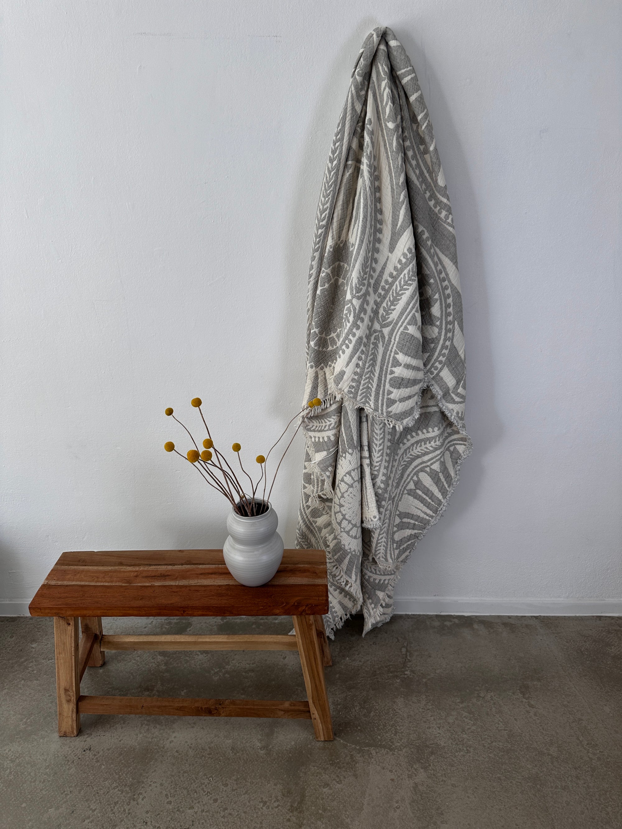 Die CALMA Musselin Decke von LEY! in Hellgrau (2,20 x 2,30 m) hängt im Boho-Stil an einer weißen Wand über einer Holzbank. Auf der Bank steht eine weiße Vase mit gelben Billy Ball Blumen auf glattem Betonboden.