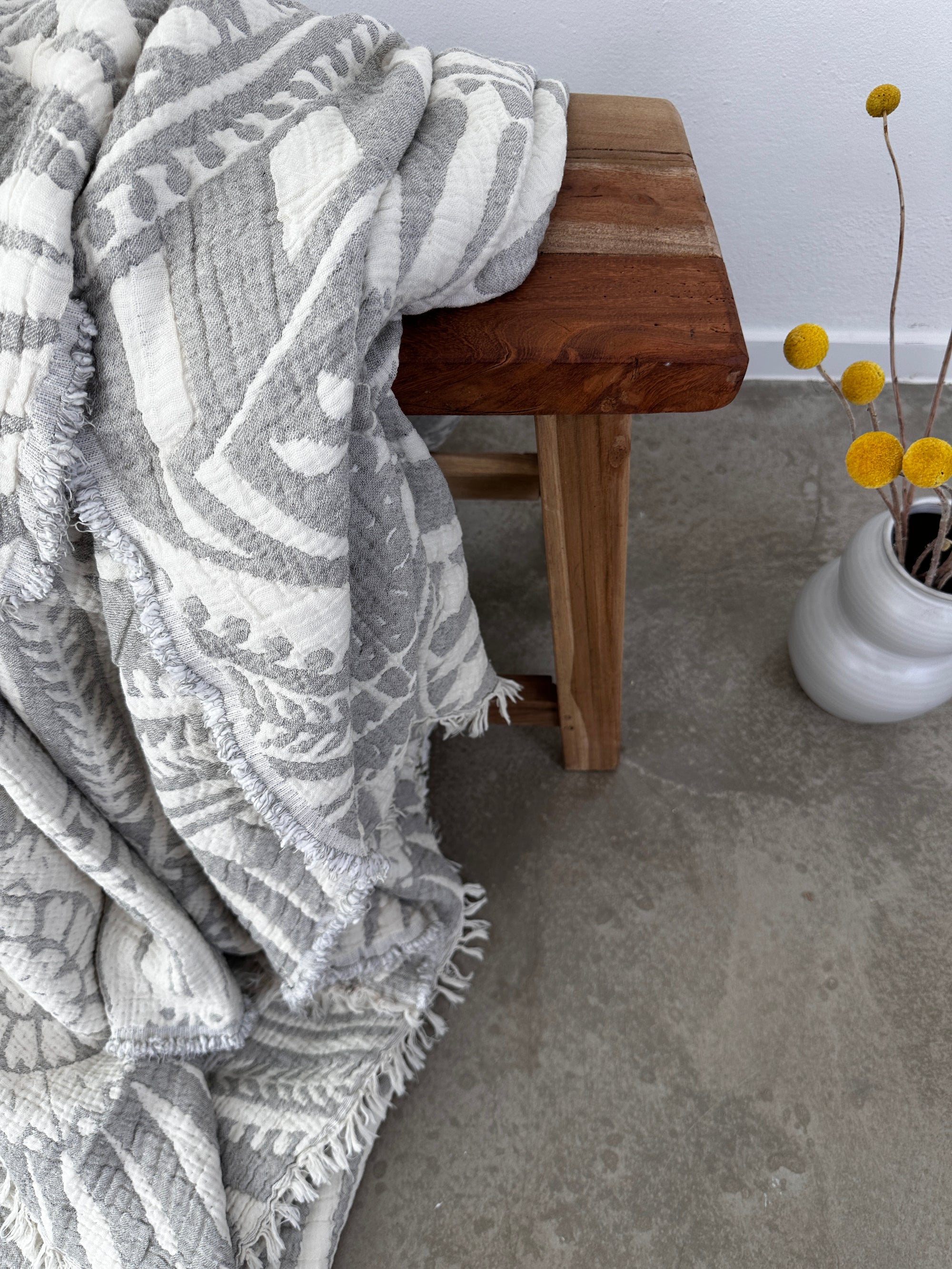 Die CALMA Musselin Decke von LEY! in Hellgrau (2,20 x 2,30 m), weich und atmungsaktiv, drapiert sich über einer Holzbank mit Fransenkanten für Boho-Flair neben einer weißen Vase mit gelben Billy Balls auf grauem Betonboden.