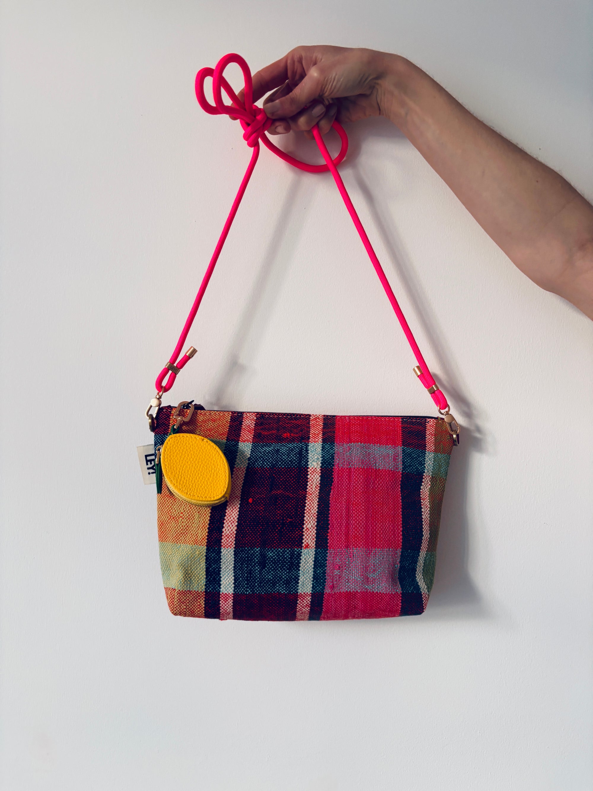 Eine Hand hält eine karierte Stoffhandtasche mit einem hellrosa Riemen und einem kleinen gelben LEY! Schlüsselanhänger - Zitrone angebracht, vor einem schlichten weißen Hintergrund.