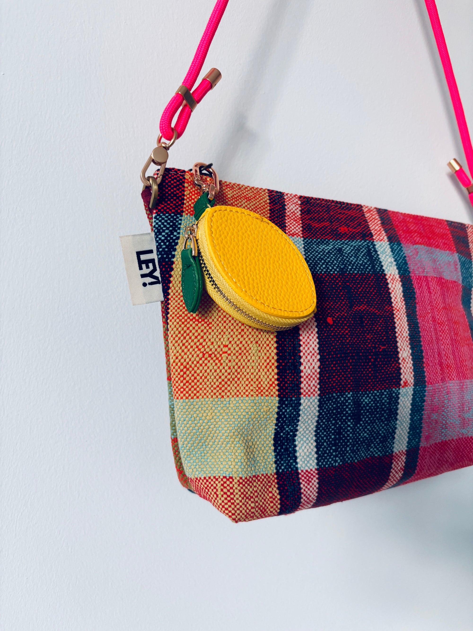Eine leuchtend karierte Handtasche mit rosa Riemen, gelbem, rundem Münzbeutel und Mini Etui Kunstleder Schlüsselanhänger - Zitrone von LEY! mit weißem "LEY!"-Schriftzug wird vor einem schlichten weißen Hintergrund präsentiert.