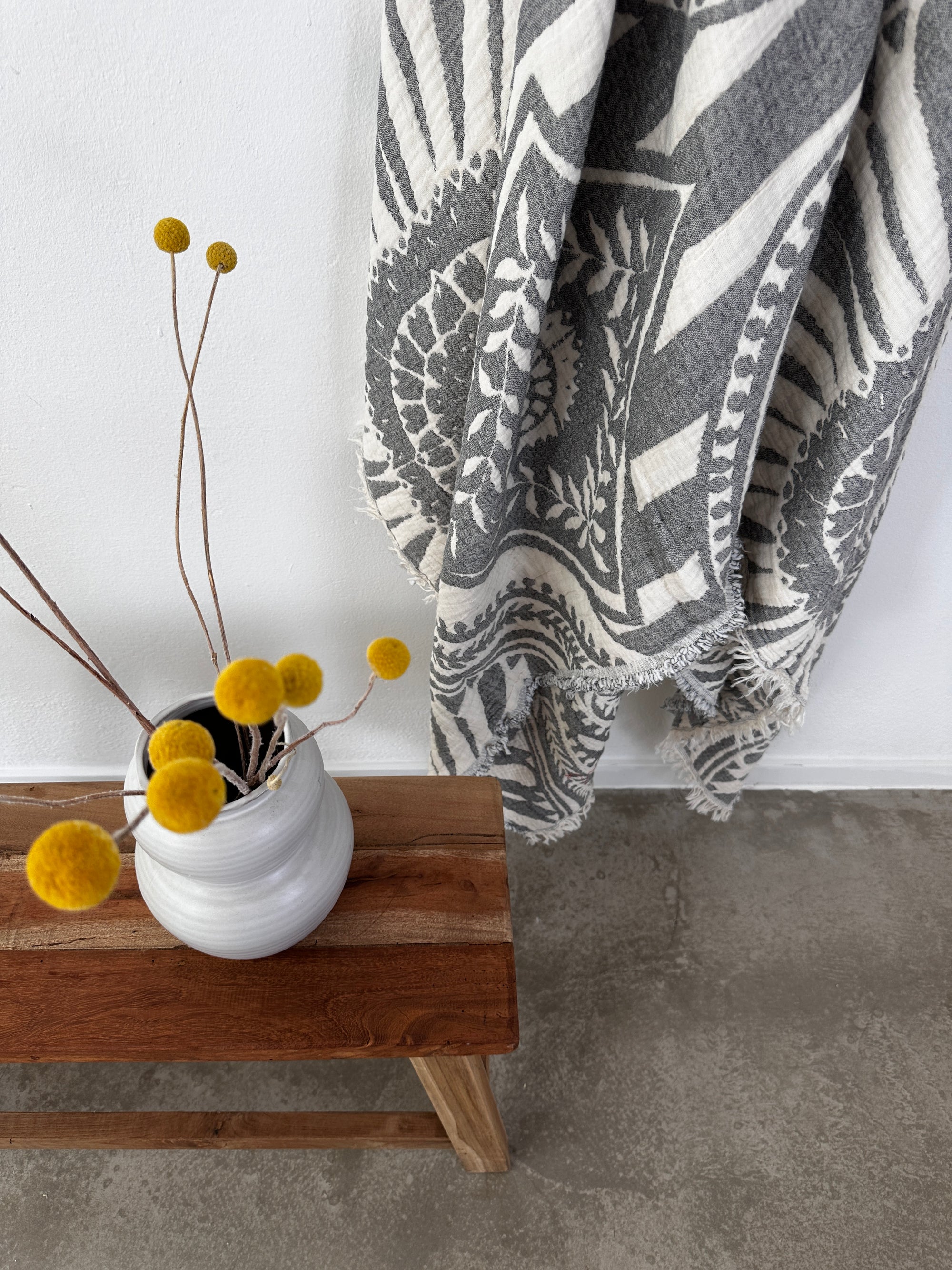 Eine weiße Vase mit gelben Billyball-Blüten steht auf einer Holzbank neben der LEY! CALMA Musselin Decke in Dunkelgrau (2,20 x 2,30 m), deren Boho-Design das graue und weiße Textil auf dem Betonboden ergänzt.