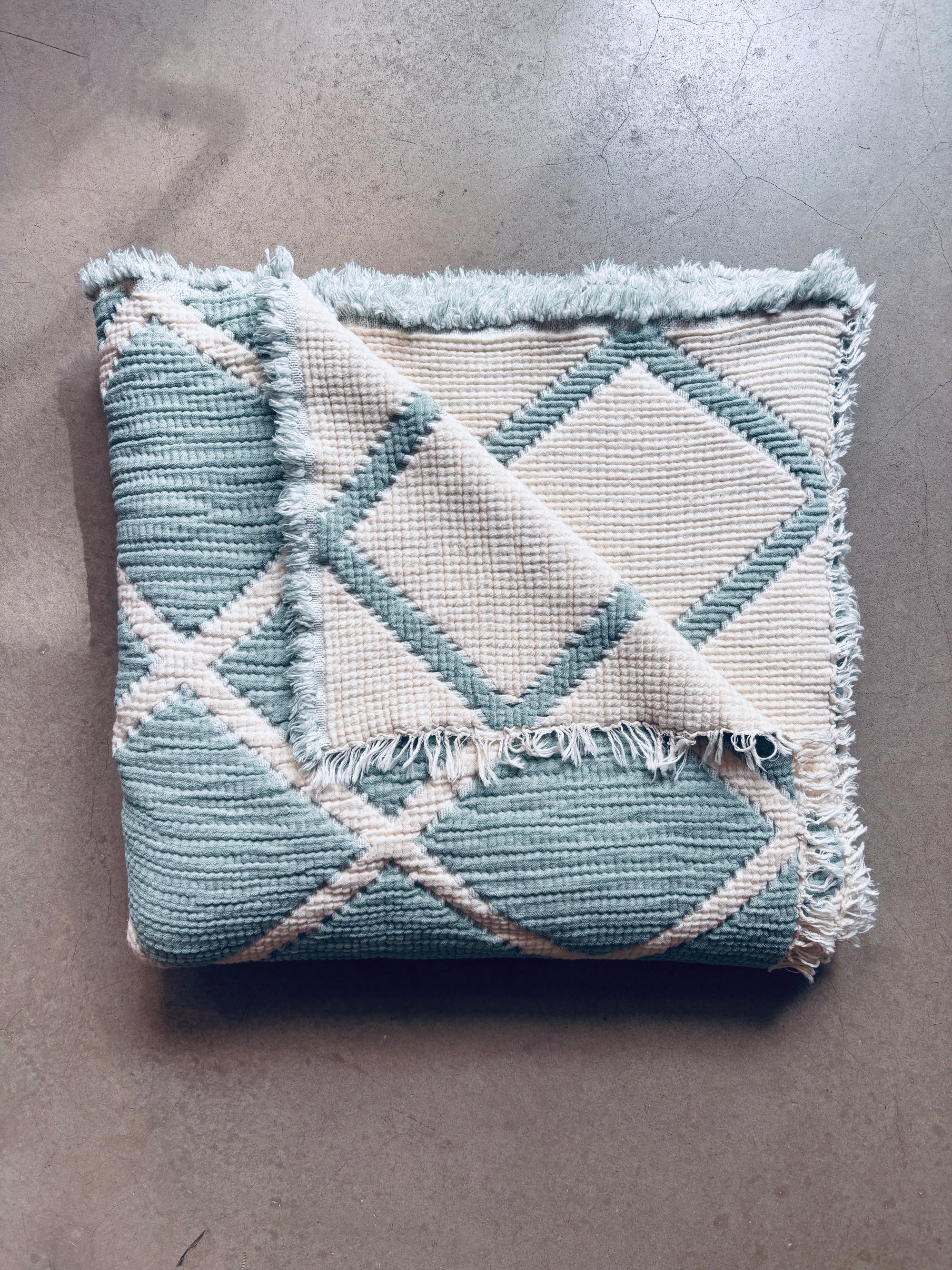 Die Musselin Decke - Salbei-Weiß (2,20 x 2,50 m) von LEY! zeigt ein salbeifarbenes und weißes Rautenmuster mit ausgefransten Kanten. Diese superweiche, hautfreundliche Decke im Boho-Design ist perfekt für helle Oberflächen.