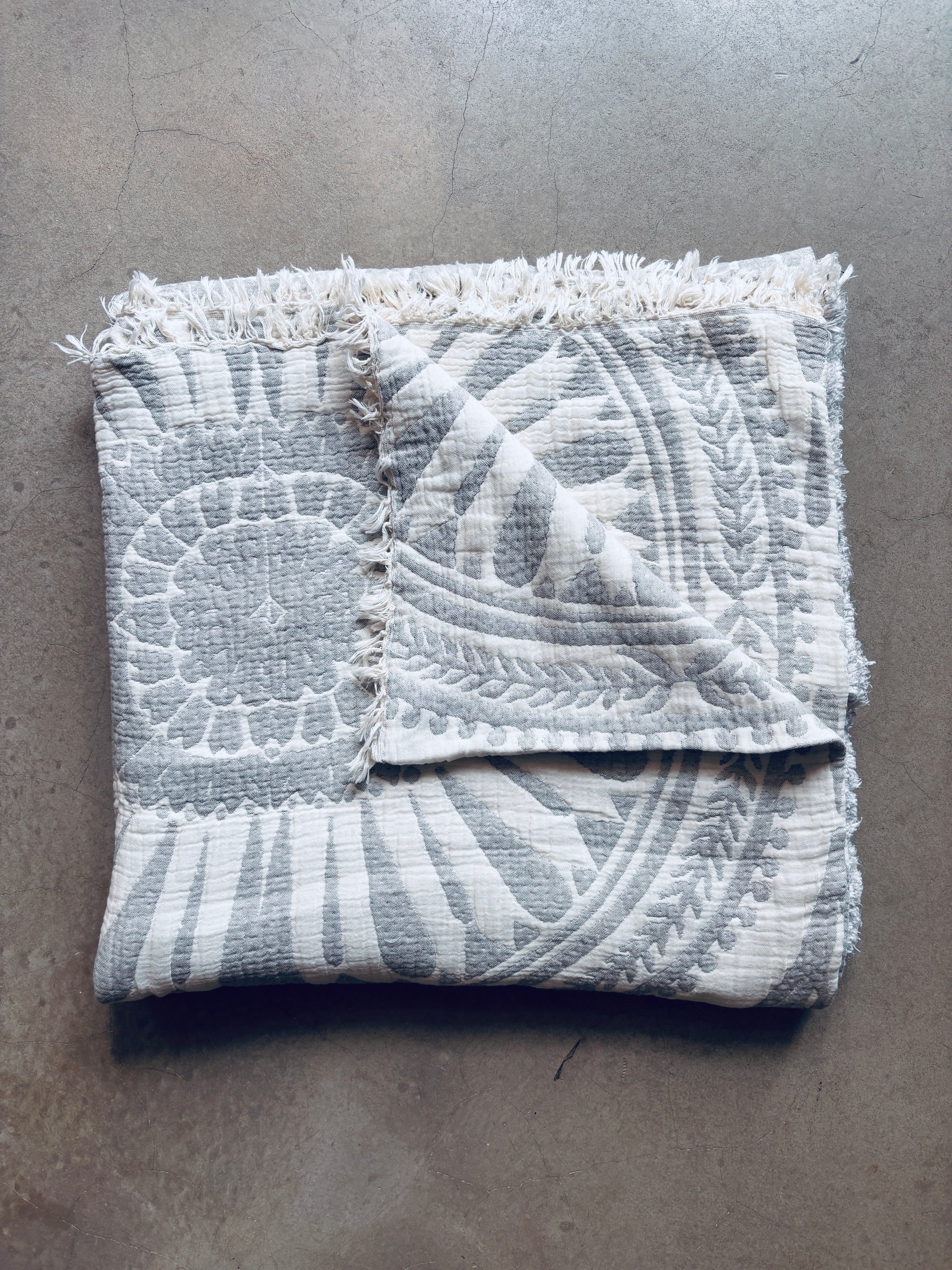 Die CALMA Musselin Decke von LEY! in Hellgrau (2,20 x 2,30 m) mit strukturiertem, gefranstem Rand und kreisförmigem Blumenmuster im Boho-Design liegt auf grauem Beton - ideal für ein gemütliches, atmungsaktives Ambiente.