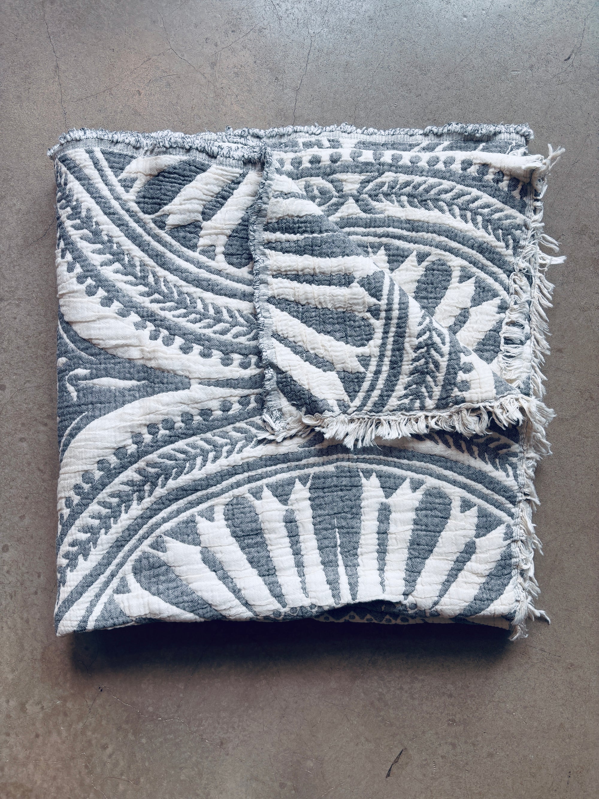 Die CALMA Musselin Decke von LEY! in dunkelgrau (2,20 x 2,30 m) mit stilvollem Boho-Design und ausgefransten Kanten ist auf einer glatten, hellgrauen Oberfläche sauber gefaltet.