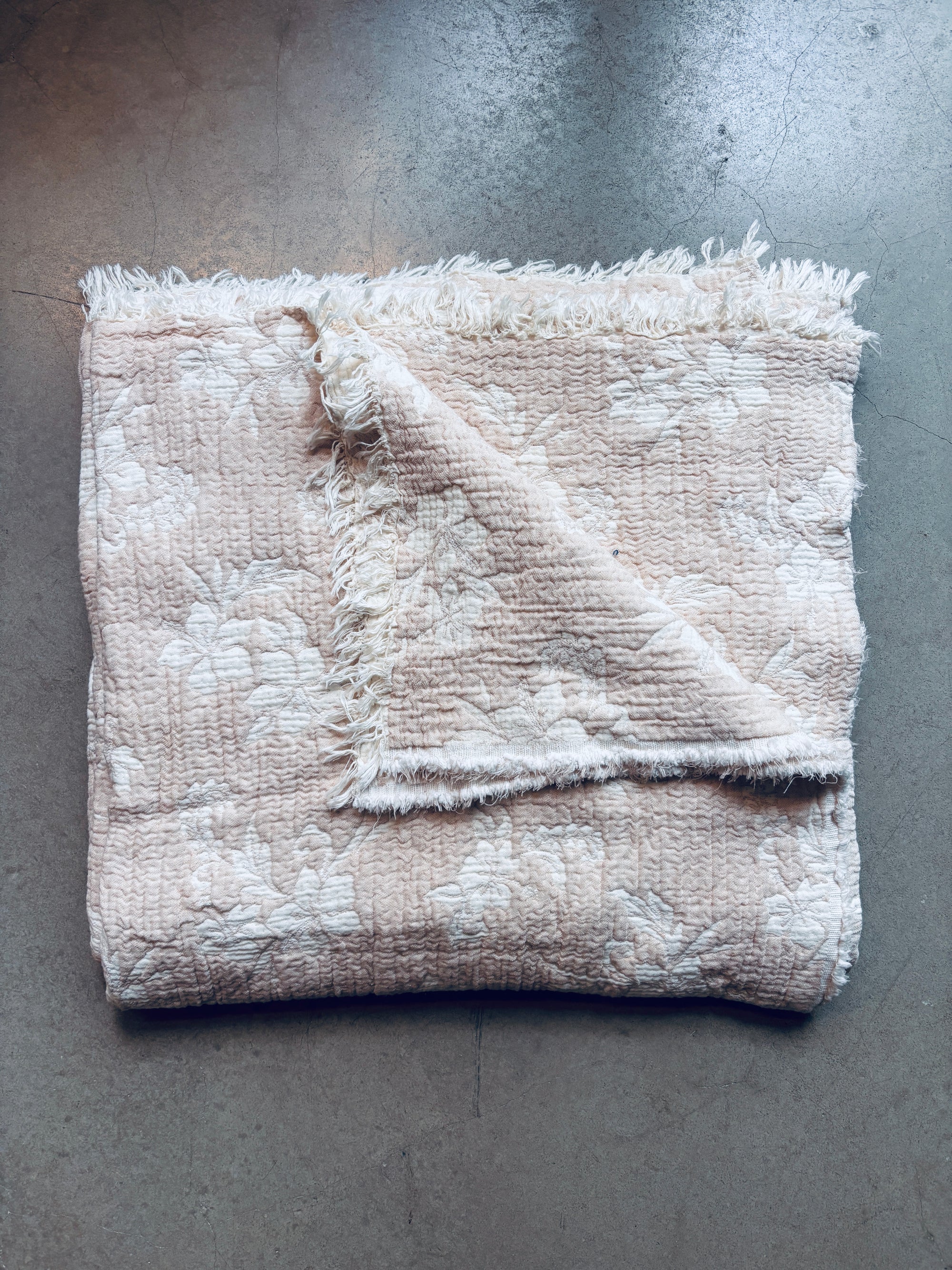 Eine gefaltete, beige-weiße Musselin Decke - ROSE by LEY! (2,20 x 2,50 m) mit dezentem Blumenmuster und ausgefransten Rändern ruht auf einer grauen Fläche, eine Ecke ist umgeschlagen, um die weiche Rückseite zu zeigen.