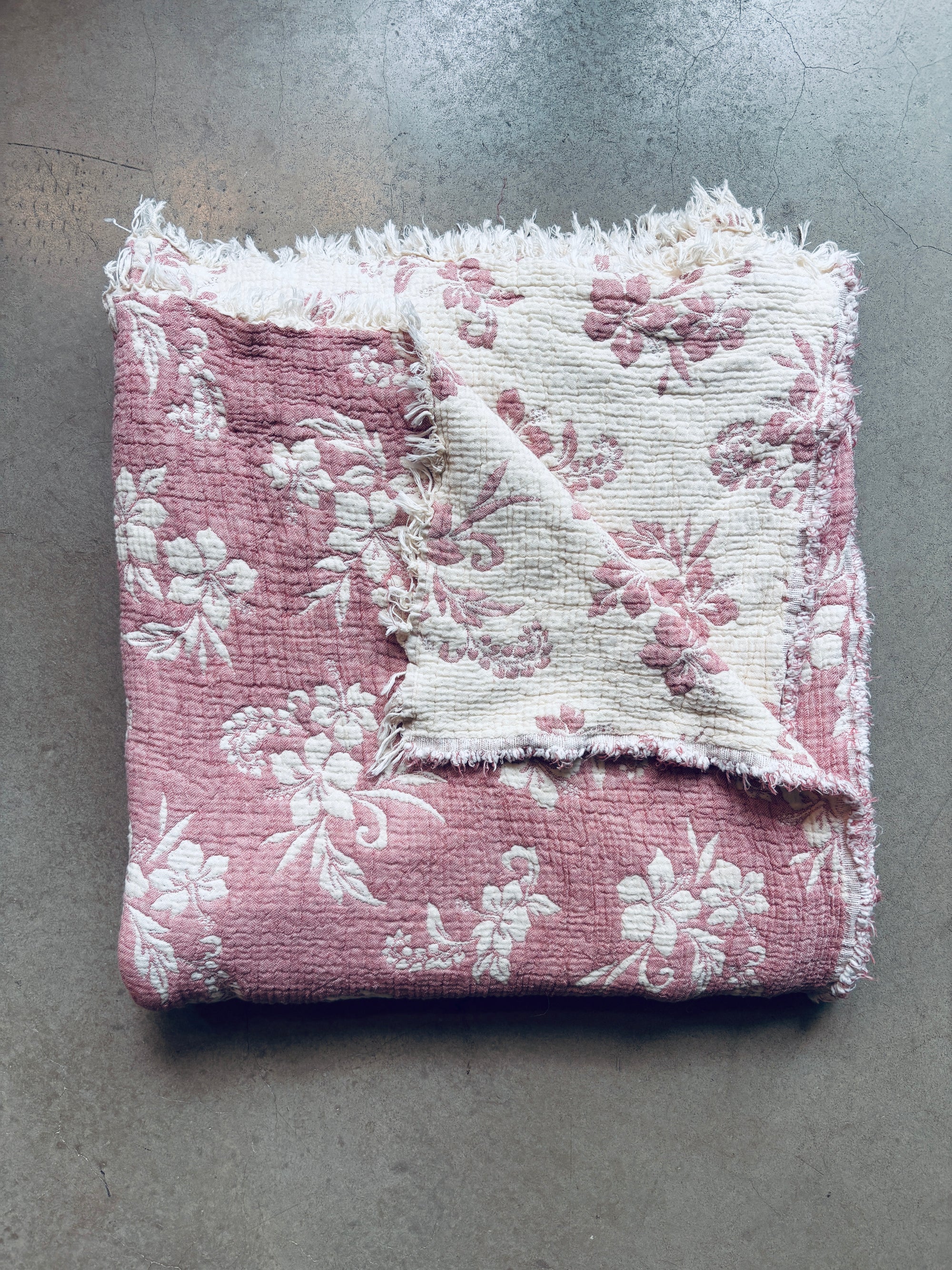 Die LEY! Musselin Decke - ROSE in Altrosa-Weiß (2,20 x 2,50 m) zeigt ein Boho-Design mit rosa und weißen Blumenmustern, ausgefransten Kanten und einer weichen, strukturierten Haptik. Abgebildet ist sie gefaltet auf einem Betonboden.