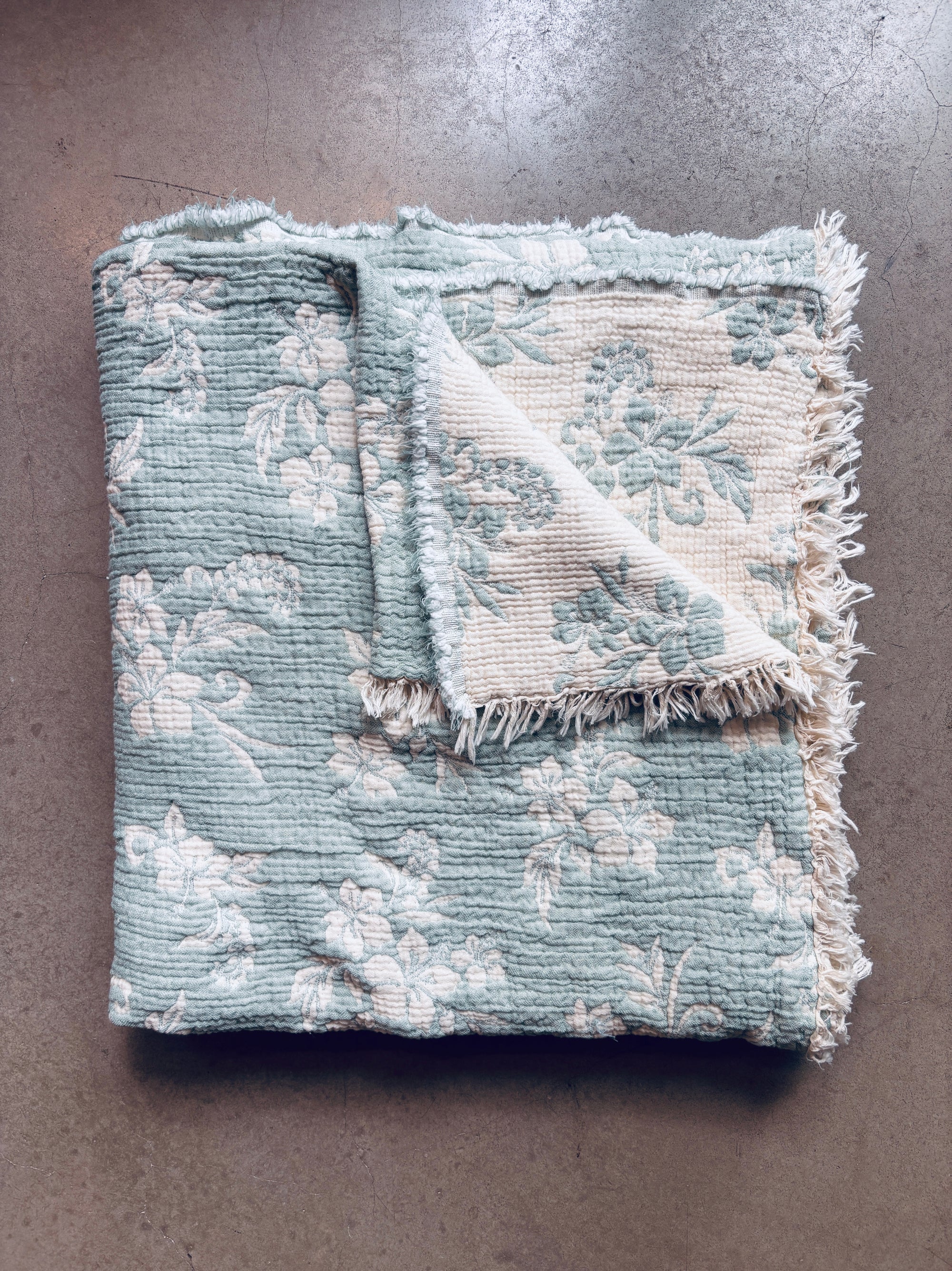 Eine gefaltete LEY! Musselin Decke - ROSE in Hellblau-Weiß (2,20 x 2,50 m) liegt auf einer grauen Fläche. Eine Ecke ist umgeschlagen und offenbart sanftere Töne und Fransen an den Kanten für gemütliches Boho-Design-Flair als kuschelige Wohndecke.