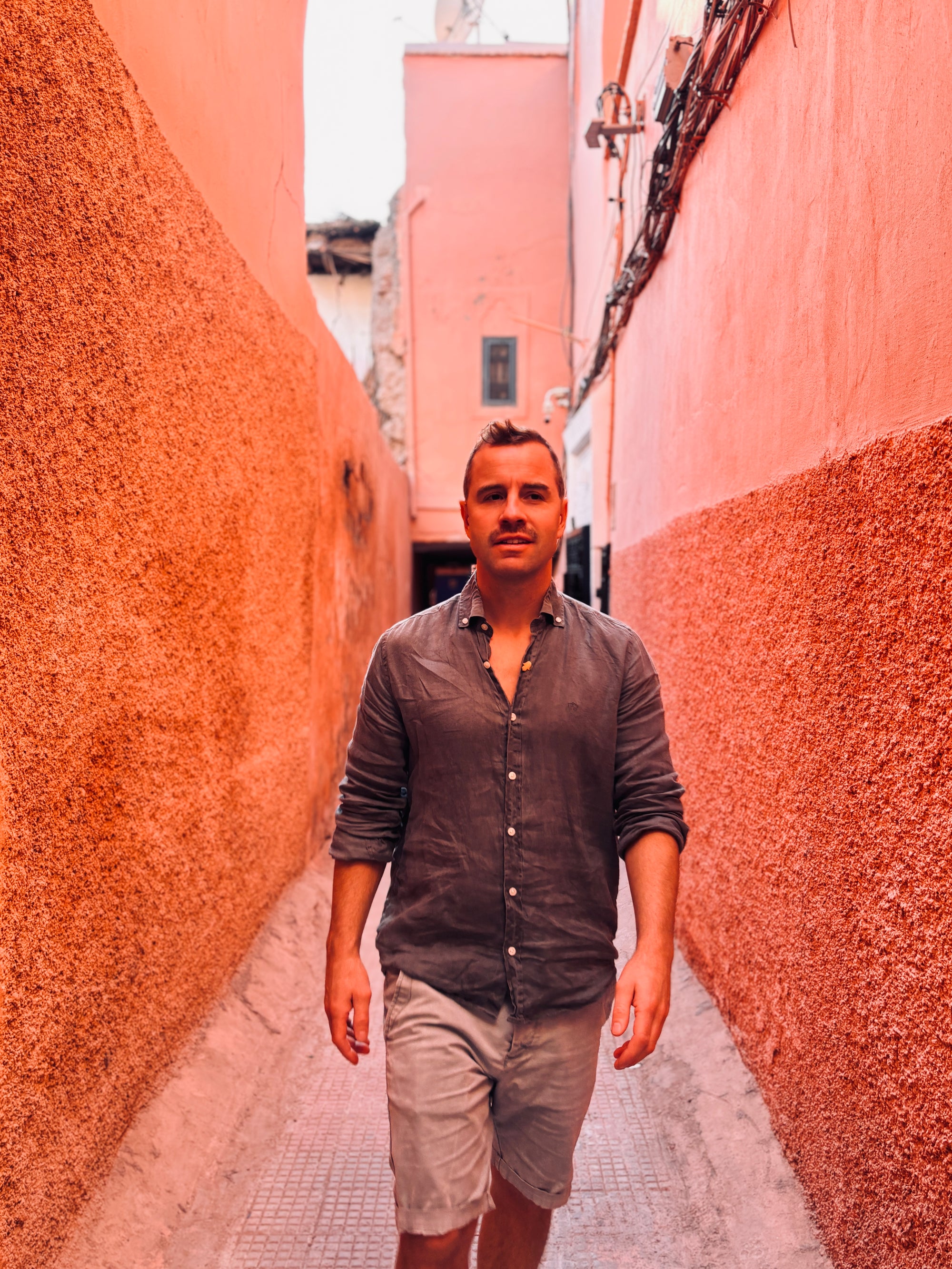 Ein Mann in einem dunklen Button-up-Hemd und hellen Shorts geht durch eine enge, leuchtend pink-orangefarbene Gasse mit strukturierten Wänden und schaut selbstbewusst nach vorne.