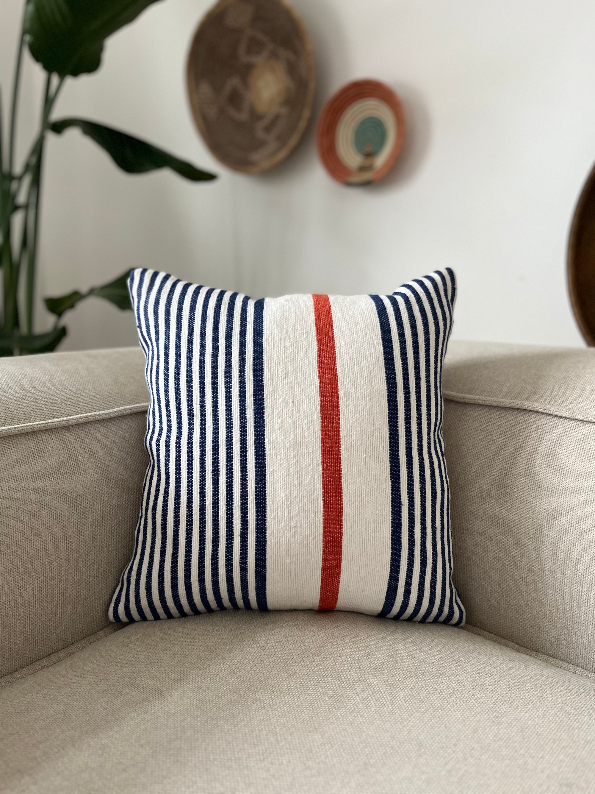 Das handgefertigte Kissen - Parosa 40x40 von LEY! mit blauen und weißen Streifen sowie rotem Akzent schmückt einen beigen Sessel, während im Hintergrund geflochtene Körbe an einer weißen Wand hängen.