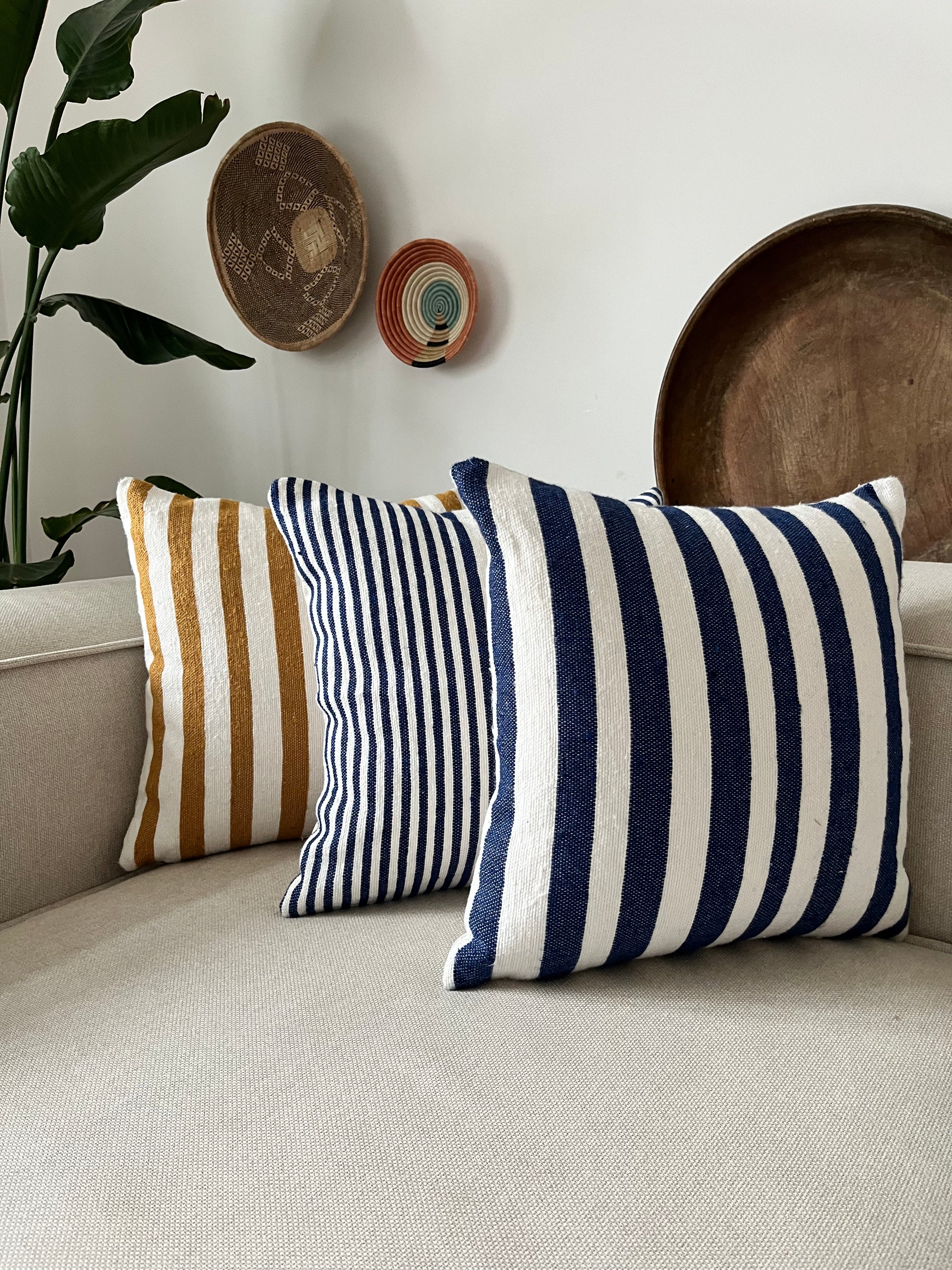 Drei gestreifte Wurfkissen, darunter das LEY! Kissen - Parosa 40x40 in Gold und Weiß, sind auf einem hellbeigen Sofa platziert. Geflochtene Körbe schmücken die weiße Wand dahinter, begleitet von einer grünen Pflanze.