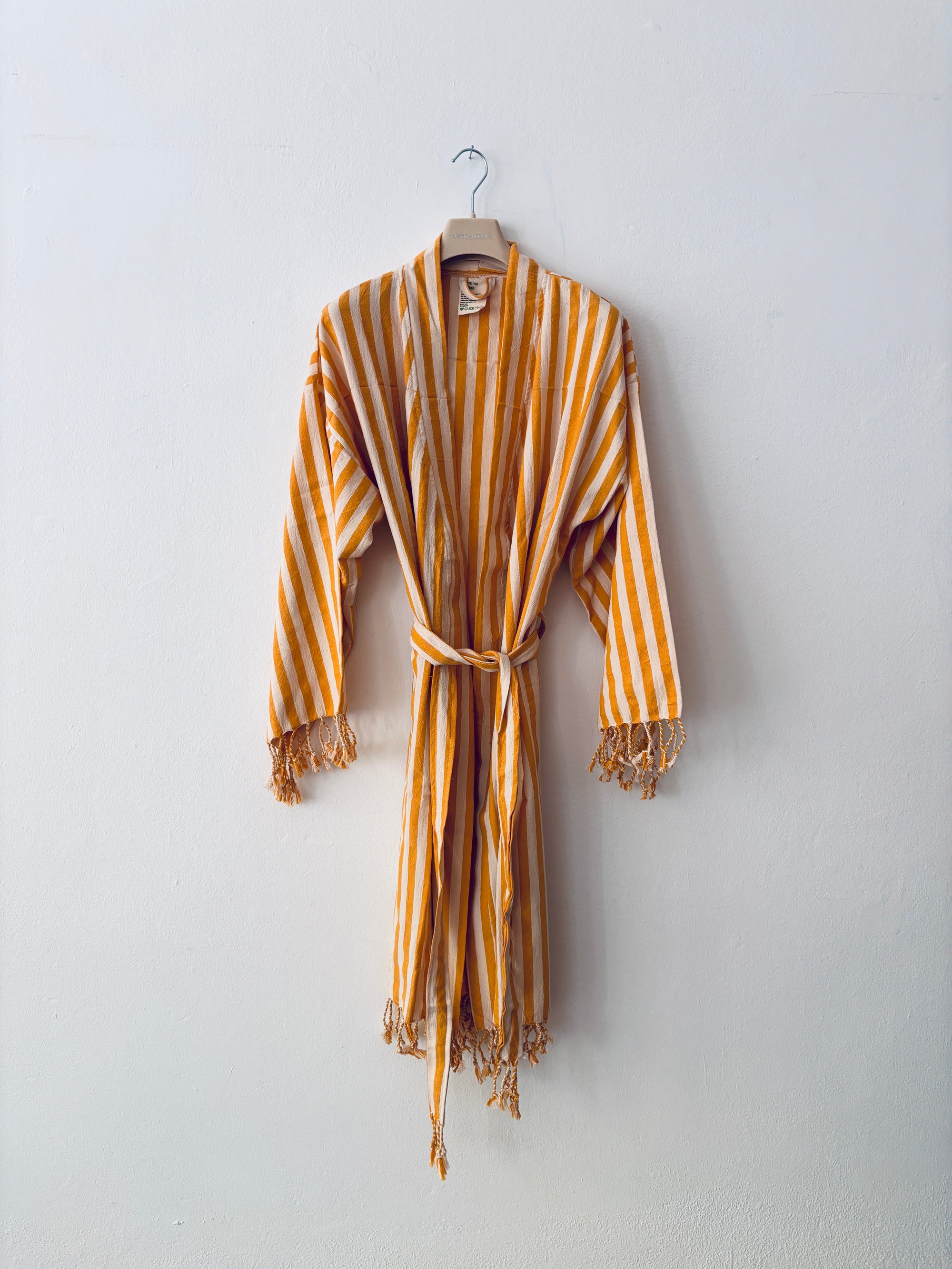Das KIMONO - MEDITERANE - GELB - WEIß von THE DANISH GIRL INTERIOR mit gelben und weißen Streifen sowie Fransen an den Ärmeln und am Saum hängt auf einem Holzbügel an einer schlichten weißen Wand.