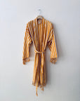 Das KIMONO - MEDITERANE - GELB - WEIß von THE DANISH GIRL INTERIOR mit gelben und weißen Streifen sowie Fransen an den Ärmeln und am Saum hängt auf einem Holzbügel an einer schlichten weißen Wand.