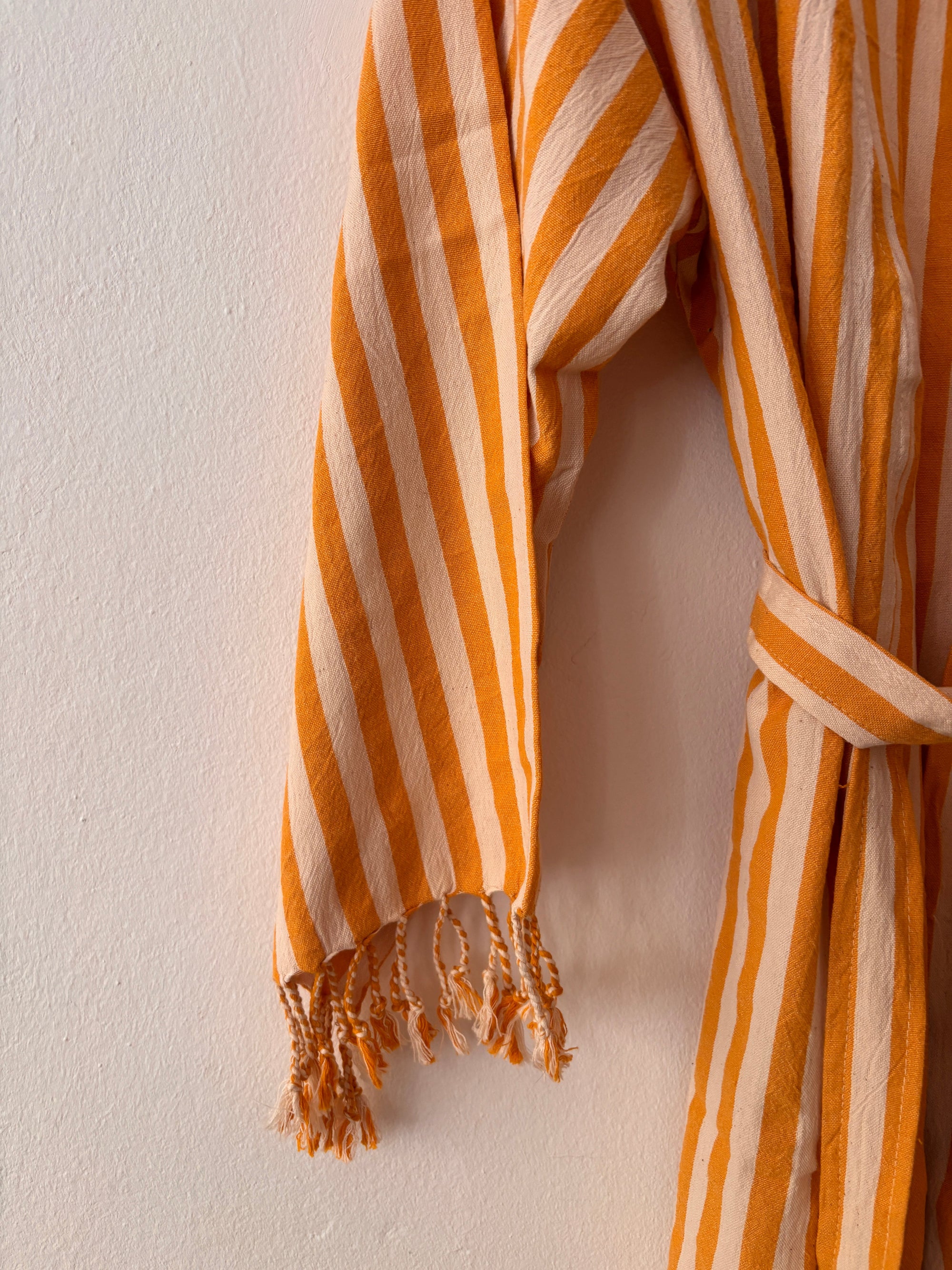 Eine Nahaufnahme des an der Wand hängenden KIMONO - MEDITERANE - GELB - WEIß von THE DANISH GIRL INTERIOR mit Taillengürtel und Fransen an den Ärmeln.