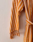 Eine Nahaufnahme des an der Wand hängenden KIMONO - MEDITERANE - GELB - WEIß von THE DANISH GIRL INTERIOR mit Taillengürtel und Fransen an den Ärmeln.